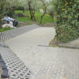 RKPOL Sp. z o.o. - Układanie ażurowej nawierzchni z kostki betonowej na podwórku. Widoczny fragment ułożonej kostki i przygotowane podłoże z geowłókniną. W tle drzewa i roślinność.