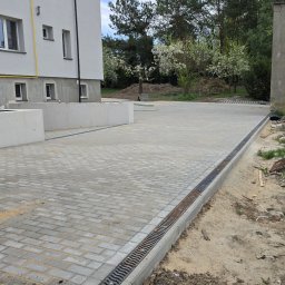 RKPOL Sp. z o.o. - Ułożona kostka brukowa na podjeździe nowego budynku z betonowymi murkami i odpływem liniowym. Widoczny fragment starego budynku i gruz.