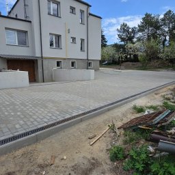 RKPOL Sp. z o.o. - Ułożona kostka brukowa na podjeździe przed budynkiem z szarą elewacją. Widoczne odwodnienie liniowe i paleta z kostką. Częściowo widoczne narzędzia i gruz.