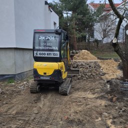 RKPOL Sp. z o.o. - Żółta minikoparka Yanmar z logo RKPOL na tle budynku i hałdy ziemi. Ślady gąsienic na błotnistym terenie. Widoczne elementy konstrukcyjne budynku i drzewa.