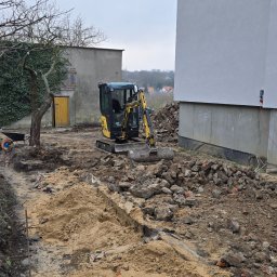 RKPOL Sp. z o.o. - Żółta mini koparka na placu budowy, przygotowuje teren pod budowę. Widoczne fundamenty budynku, gruz i wykopany rów w ziemi. Na drugim planie budynek gospodarczy.