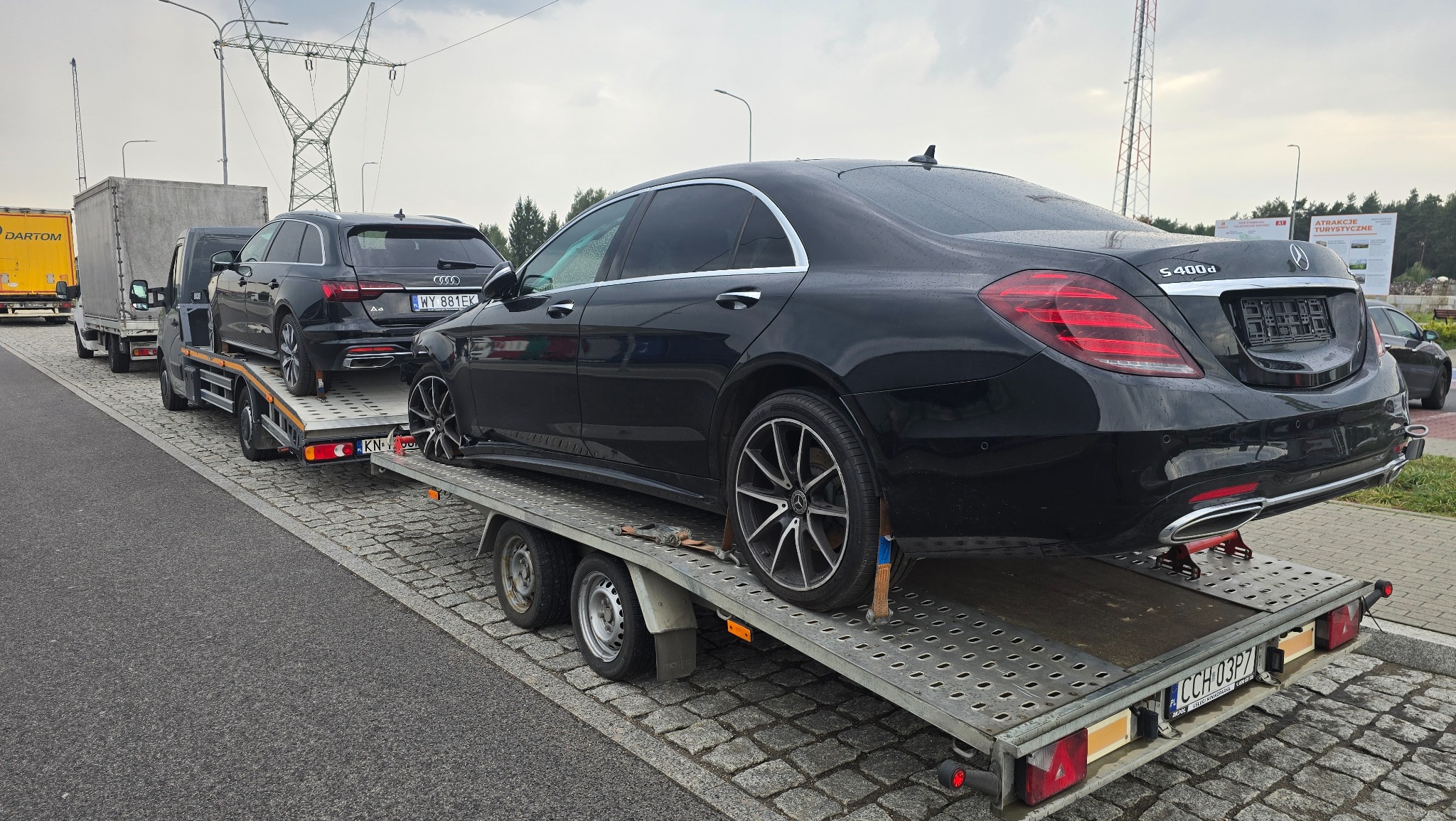 Czarny Mercedes S400d na niskopodwoziowej lawecie, zabezpieczony pasami transportowymi, w tle drugi samochód Audi A4 i ciężarówka Dartom. Transport aut.