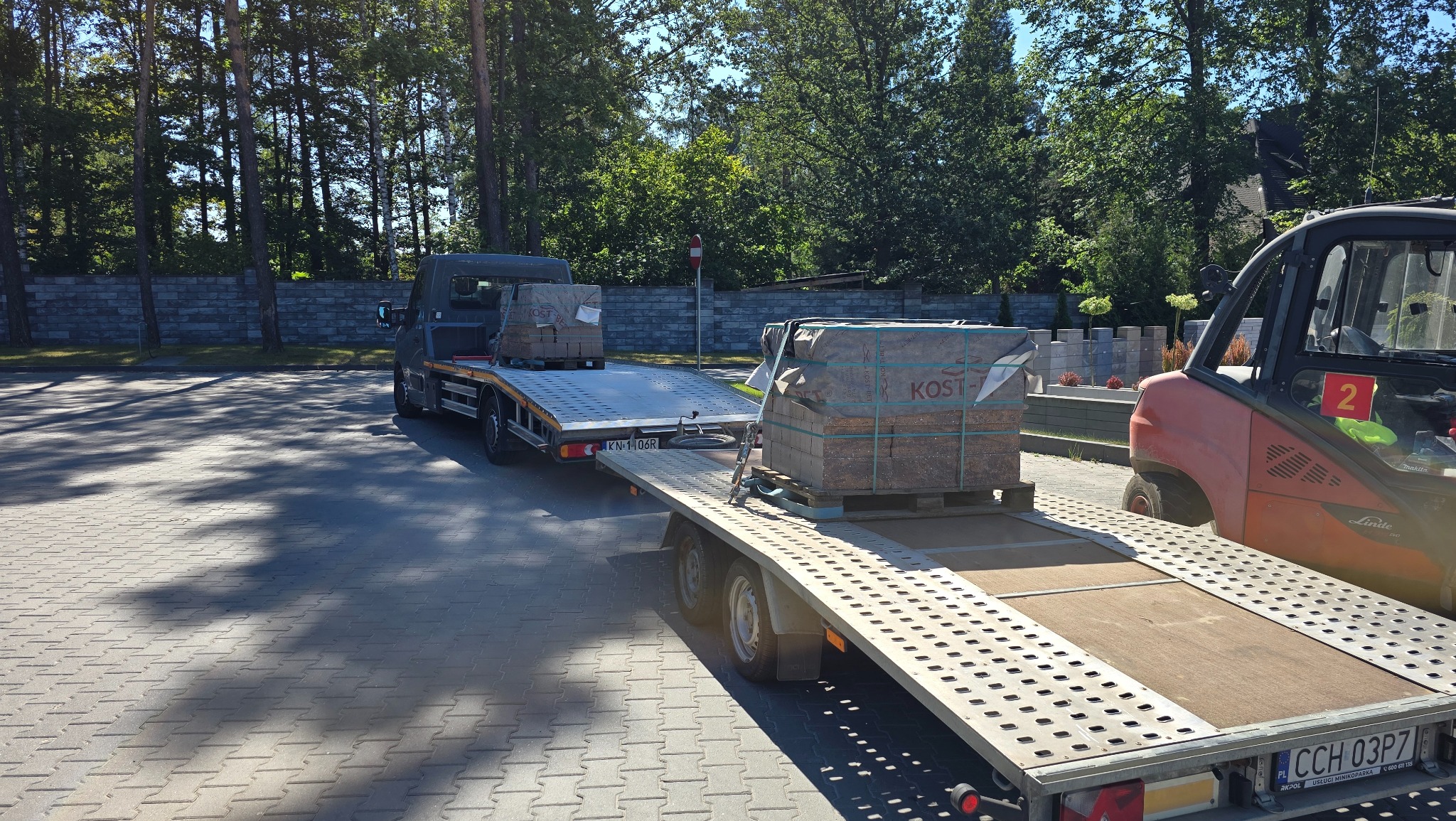 Laweta z kostką brukową na przyczepie, widoczny wózek widłowy. Kostka marki Kost-Bet. Tło: drzewa i mur. Teren wyłożony kostką brukową.