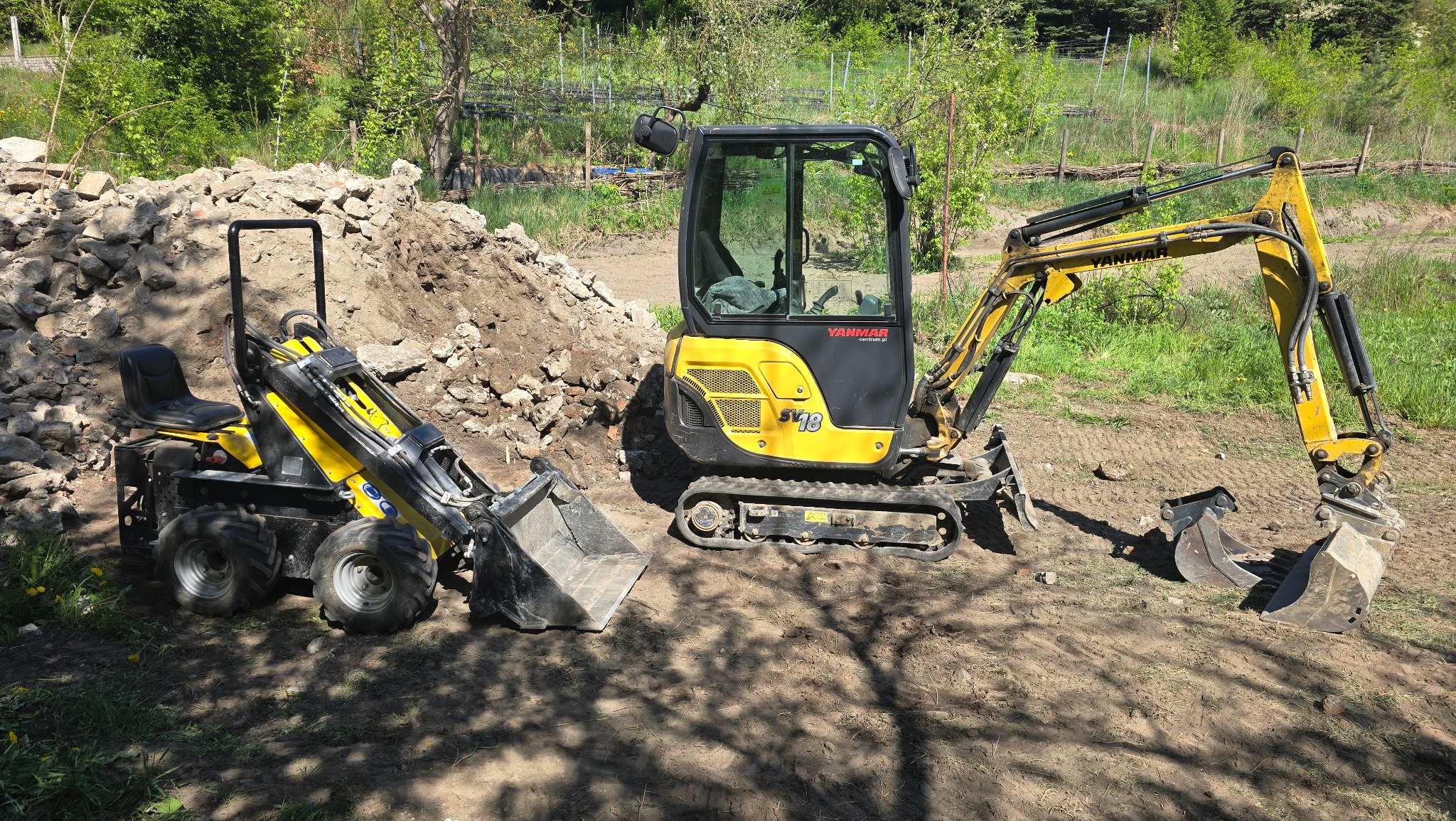 Żółty minikoparka Yanmar SV18 i minitraktor z łyżką na placu budowy, w tle hałda ziemi i kamieni. Sprzęt budowlany w naturalnym otoczeniu, w słoneczny dzień.