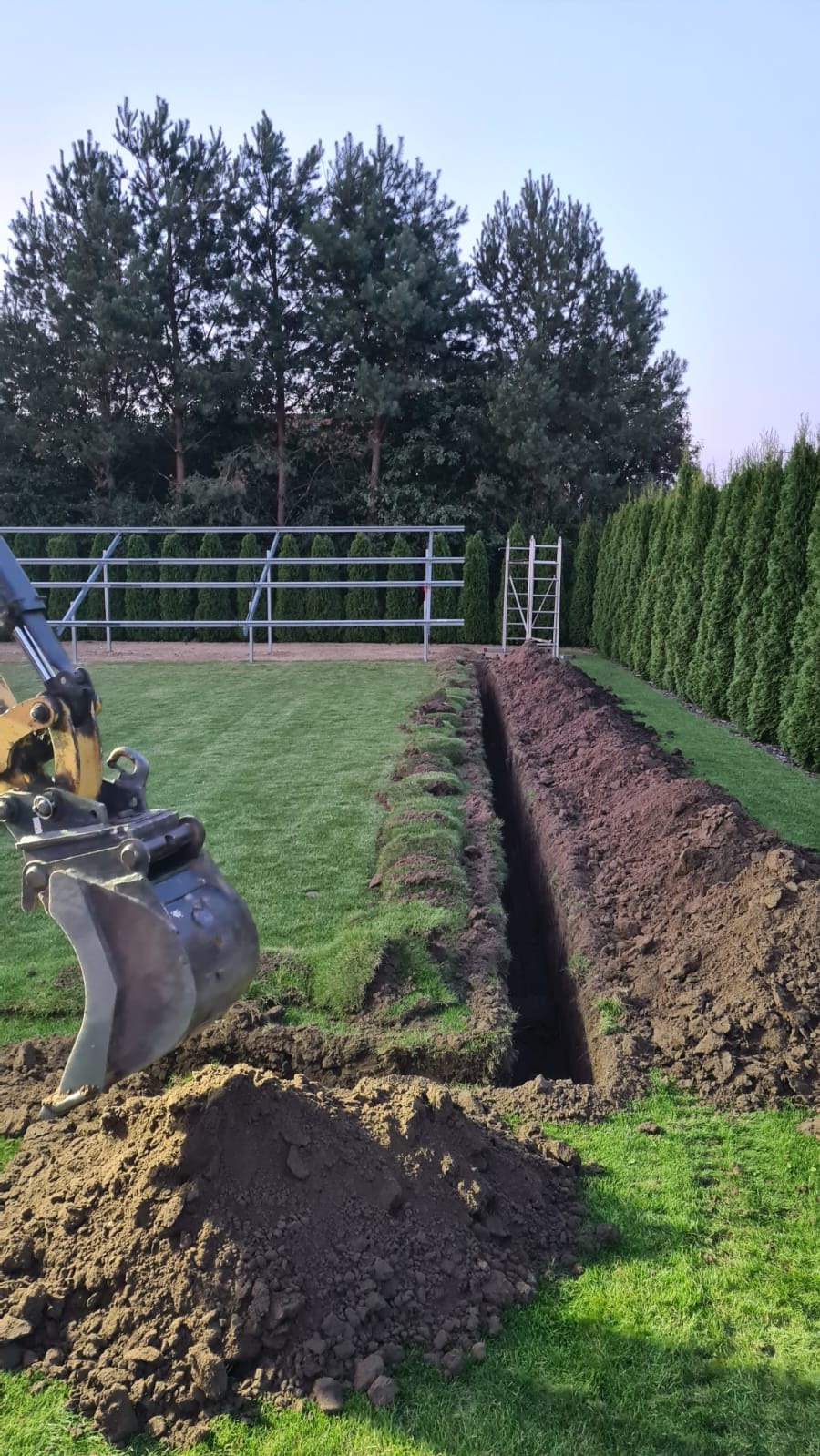 Wykopany wąski rów w trawie, obok sterta ziemi i ramię koparki. W tle rząd tuj i metalowa konstrukcja.