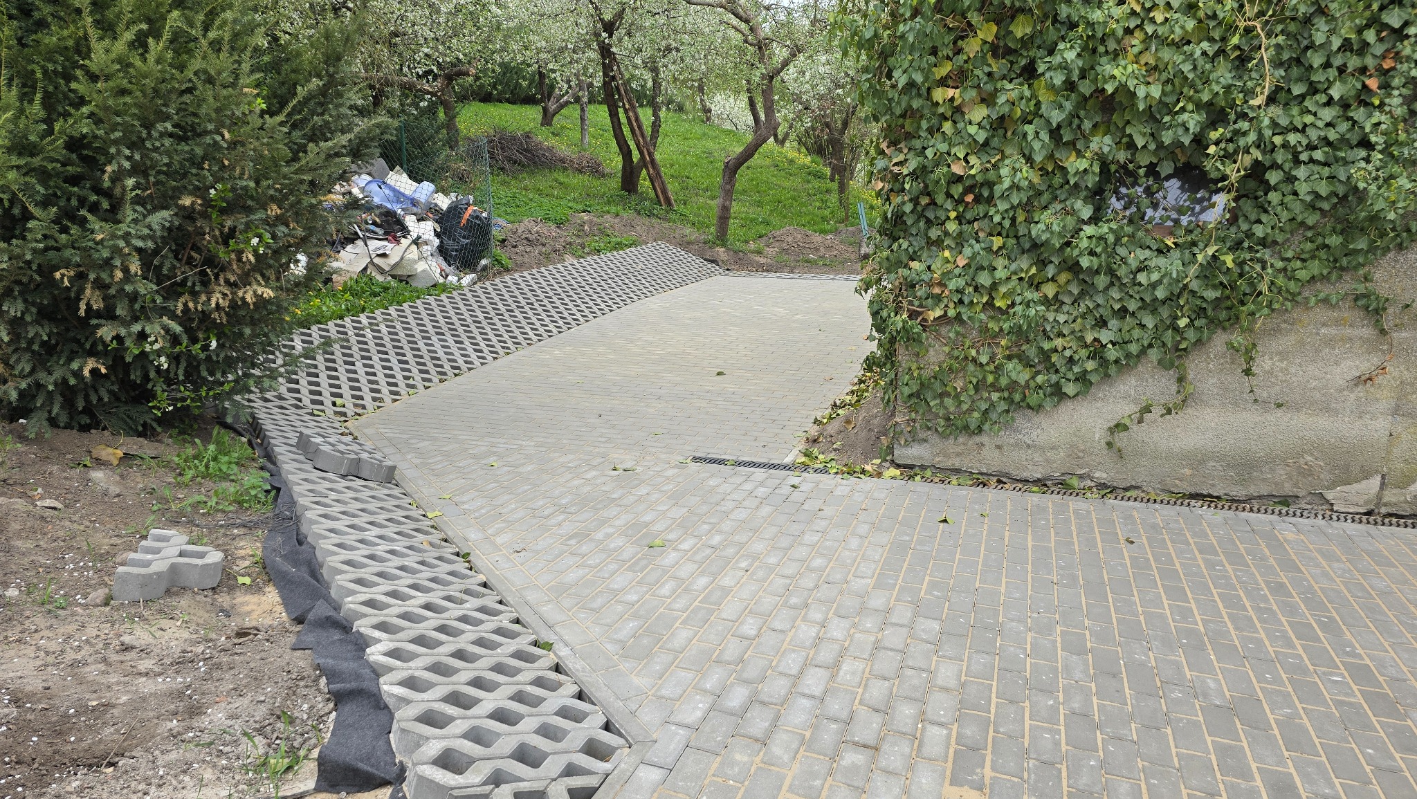 Układanie ażurowej nawierzchni z kostki betonowej na podwórku. Widoczny fragment ułożonej kostki i przygotowane podłoże z geowłókniną. W tle drzewa i roślinność.