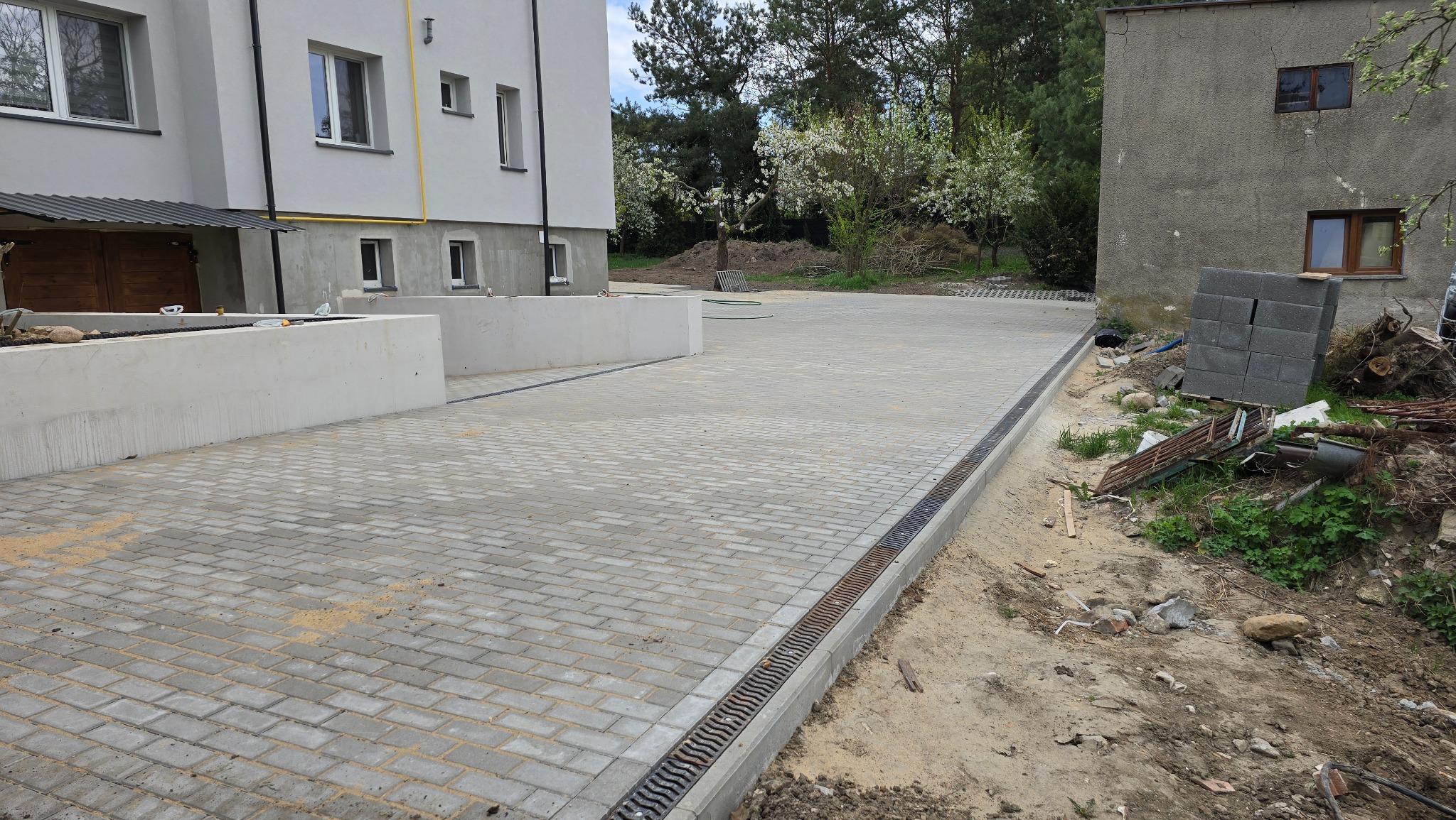 Ułożona kostka brukowa na podjeździe nowego budynku z betonowymi murkami i odpływem liniowym. Widoczny fragment starego budynku i gruz.
