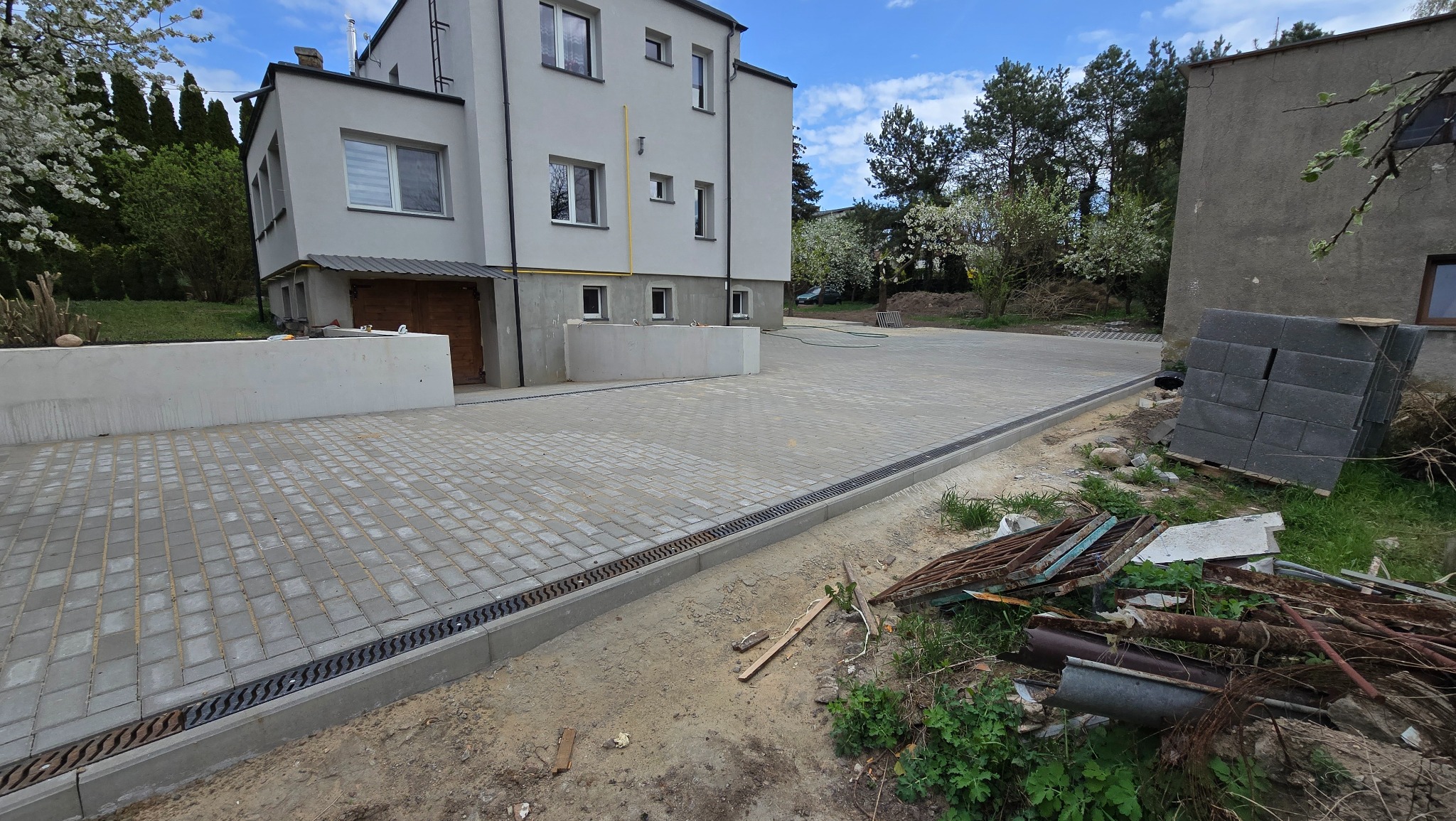 Ułożona kostka brukowa na podjeździe przed budynkiem z szarą elewacją. Widoczne odwodnienie liniowe i paleta z kostką. Częściowo widoczne narzędzia i gruz.