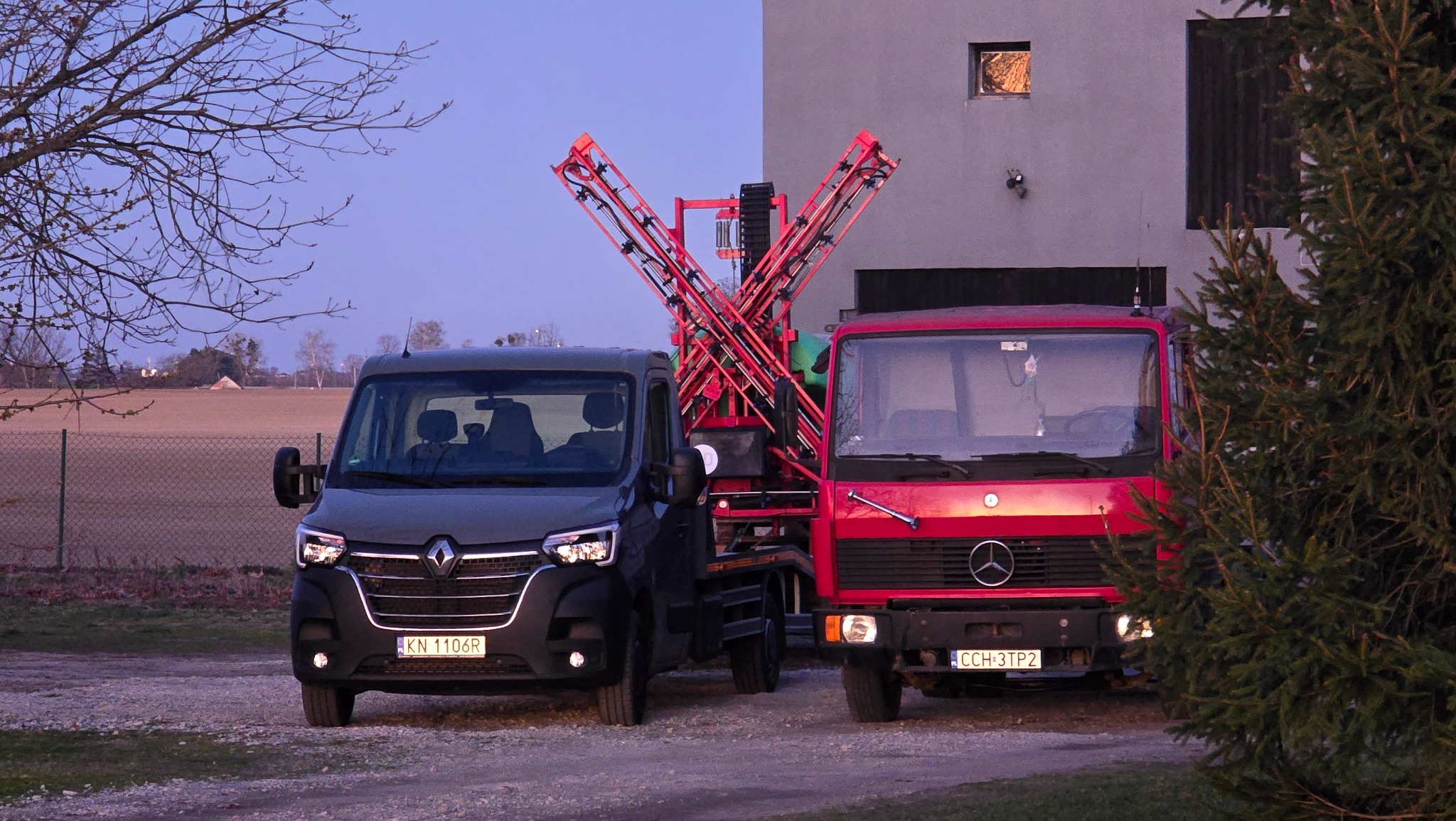 Dwa samochody: szary Renault Master i czerwony Mercedes, z zamontowanym opryskiwaczem polowym, stoją na żwirowej drodze, na tle budynku i pola.
