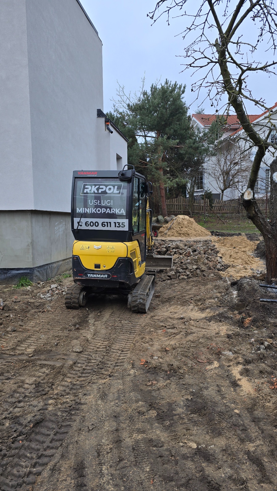 Żółta minikoparka Yanmar z logo RKPOL na tle budynku i hałdy ziemi. Ślady gąsienic na błotnistym terenie. Widoczne elementy konstrukcyjne budynku i drzewa.