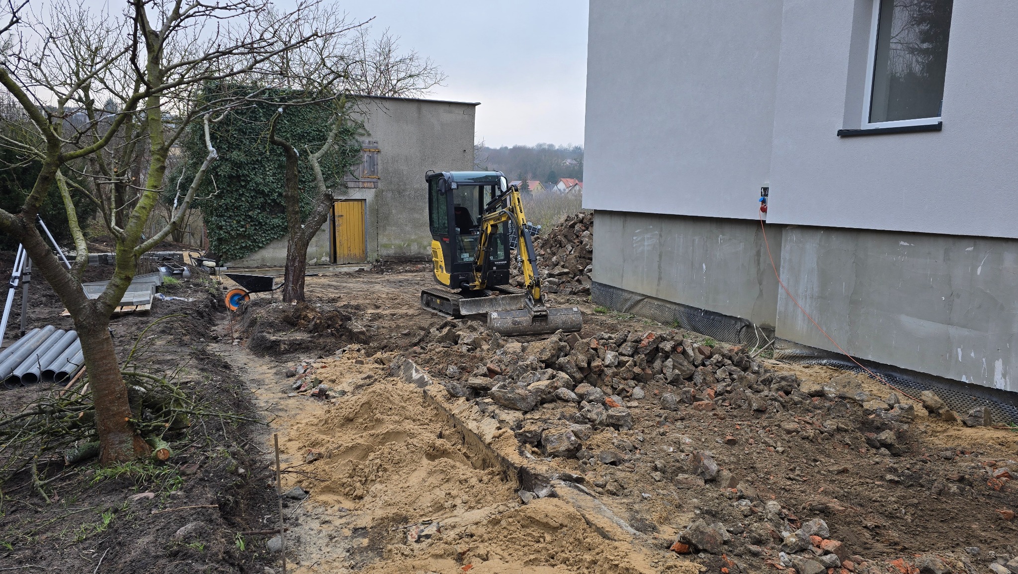 Żółta mini koparka na placu budowy, przygotowuje teren pod budowę. Widoczne fundamenty budynku, gruz i wykopany rów w ziemi. Na drugim planie budynek gospodarczy.