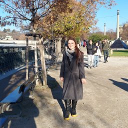 Pracownia Krawiecka - Atelier Marta Sierny-Kroczek - Kobieta w szarym płaszczu i czarnych botkach na tle parku w słoneczny dzień. Jesienne drzewo z brązowymi liśćmi i alejka z ławkami.