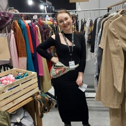 Pracownia Krawiecka - Atelier Marta Sierny-Kroczek - Uśmiechnięta kobieta w czarnej sukience prezentuje się na stoisku z odzieżą, z różnorodnymi ubraniami na wieszakach i drewnianą skrzynią z akcesoriami obok.