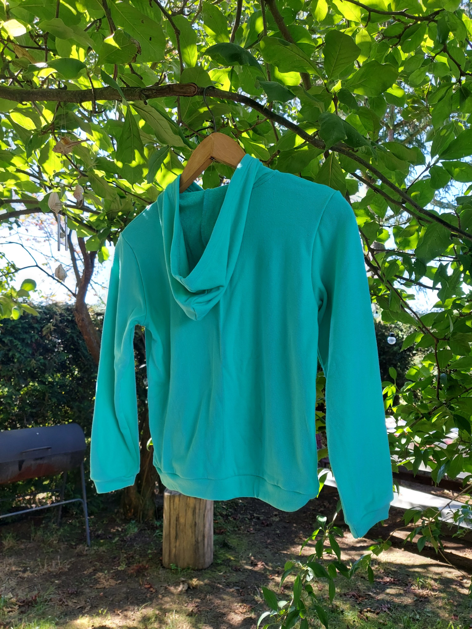 Turkusowa bluza z kapturem, wisząca na drewnianym wieszaku na tle zielonych liści drzewa i ogrodu. Widoczny grill i pień drzewa.