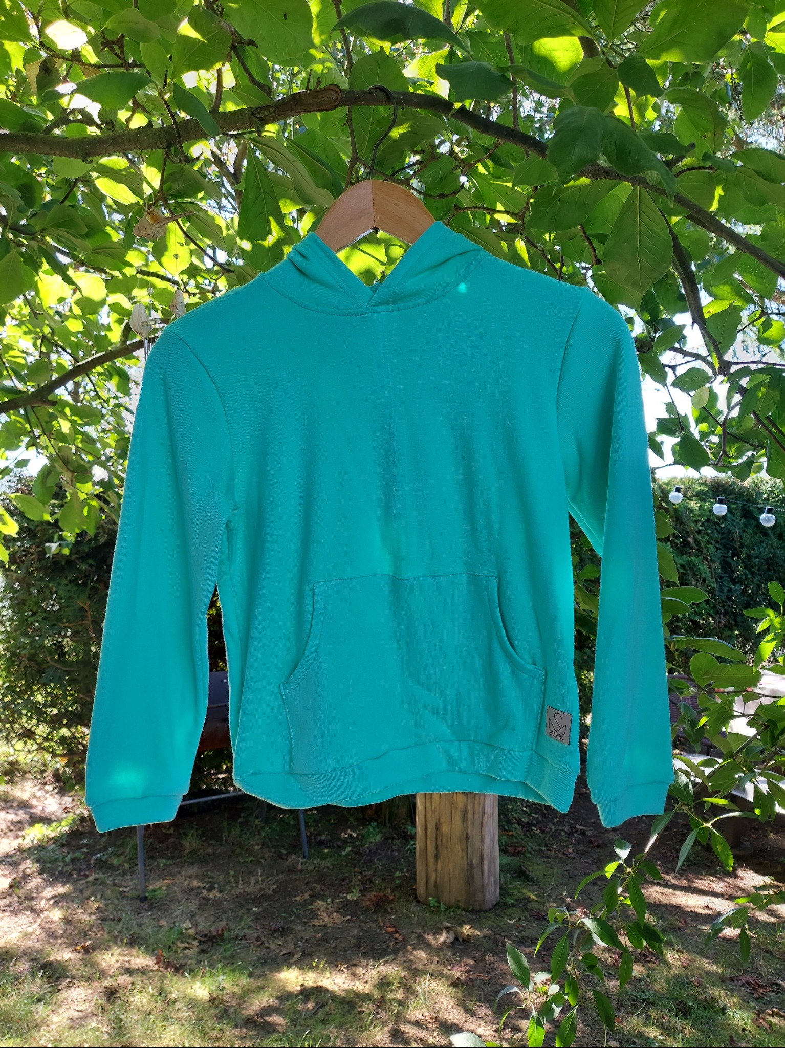 Turkusowa bluza z kapturem, zawieszona na wieszaku na tle zielonych liści i słonecznego ogrodu. Widoczny detal metki z logo marki. Casualowy styl.