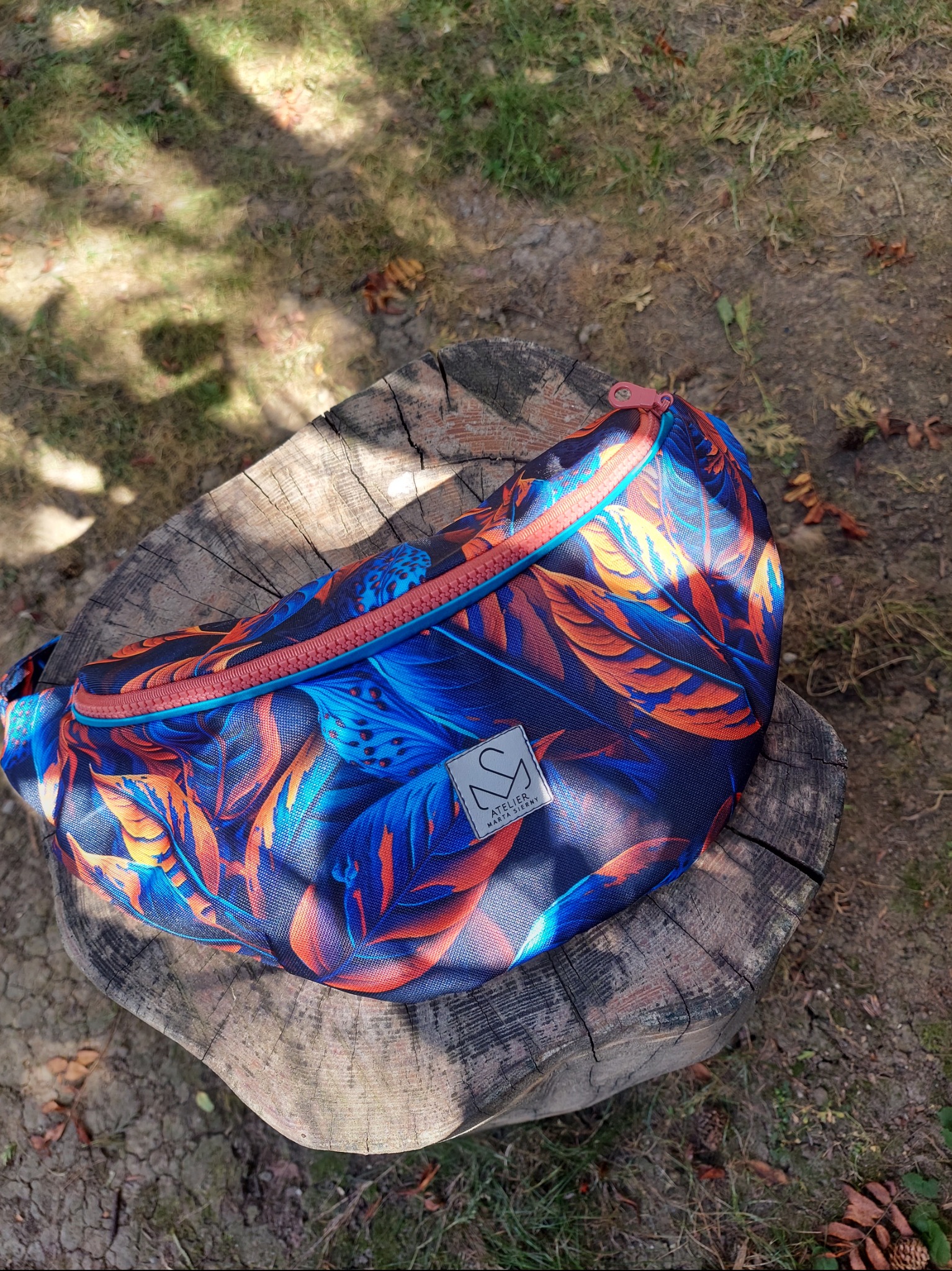 Kolorowa nerka z motywem liści na drewnianym pniu w plenerze. Widoczne logo 'Atelier Marta Cierny'. Unikalny wzór i naturalne otoczenie dodają uroku.