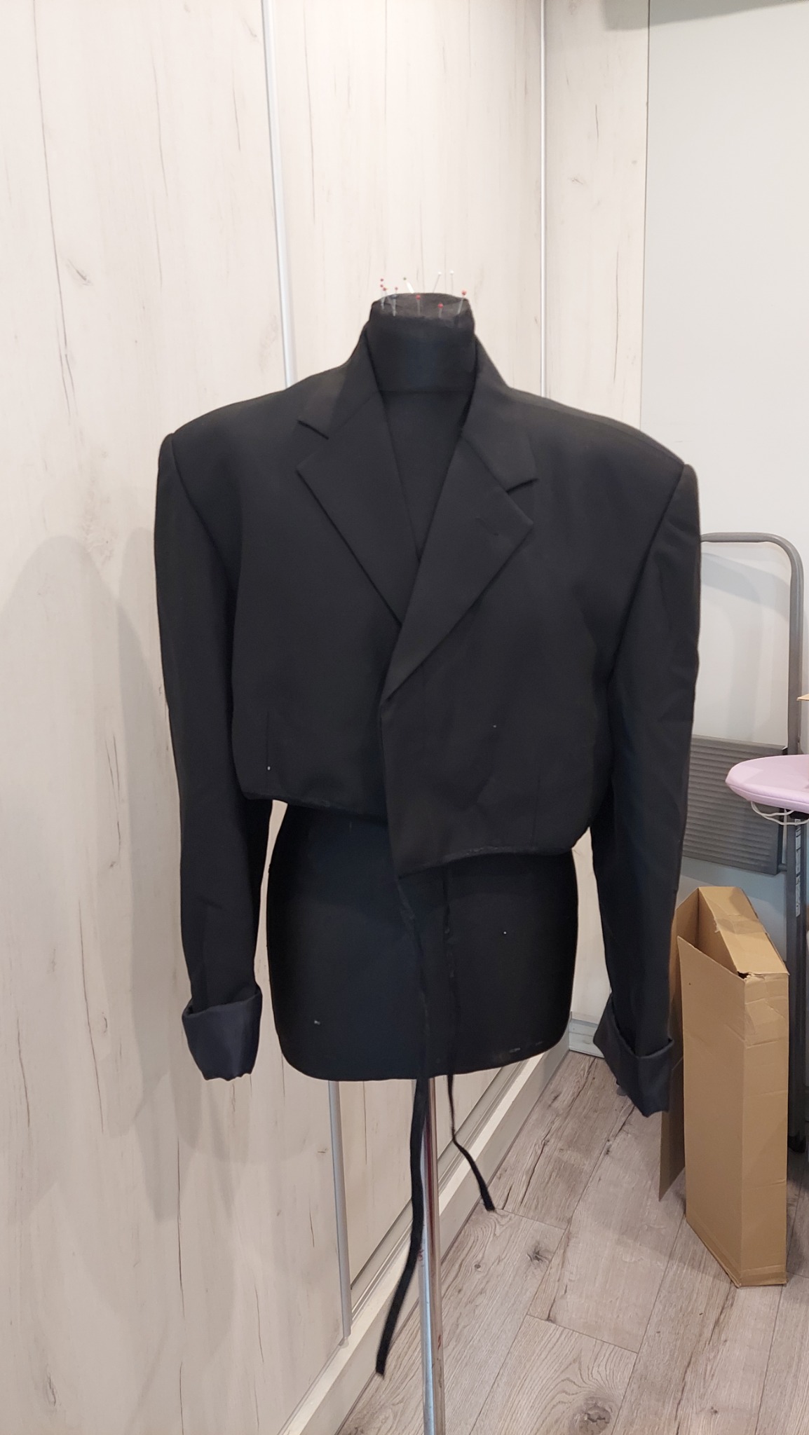 Krótki, czarny żakiet damski na manekinie krawieckim, z widocznymi szpilkami w okolicy szyi, na tle jasnej ściany z drewnopodobnym wzorem i fragmentem drabinki.