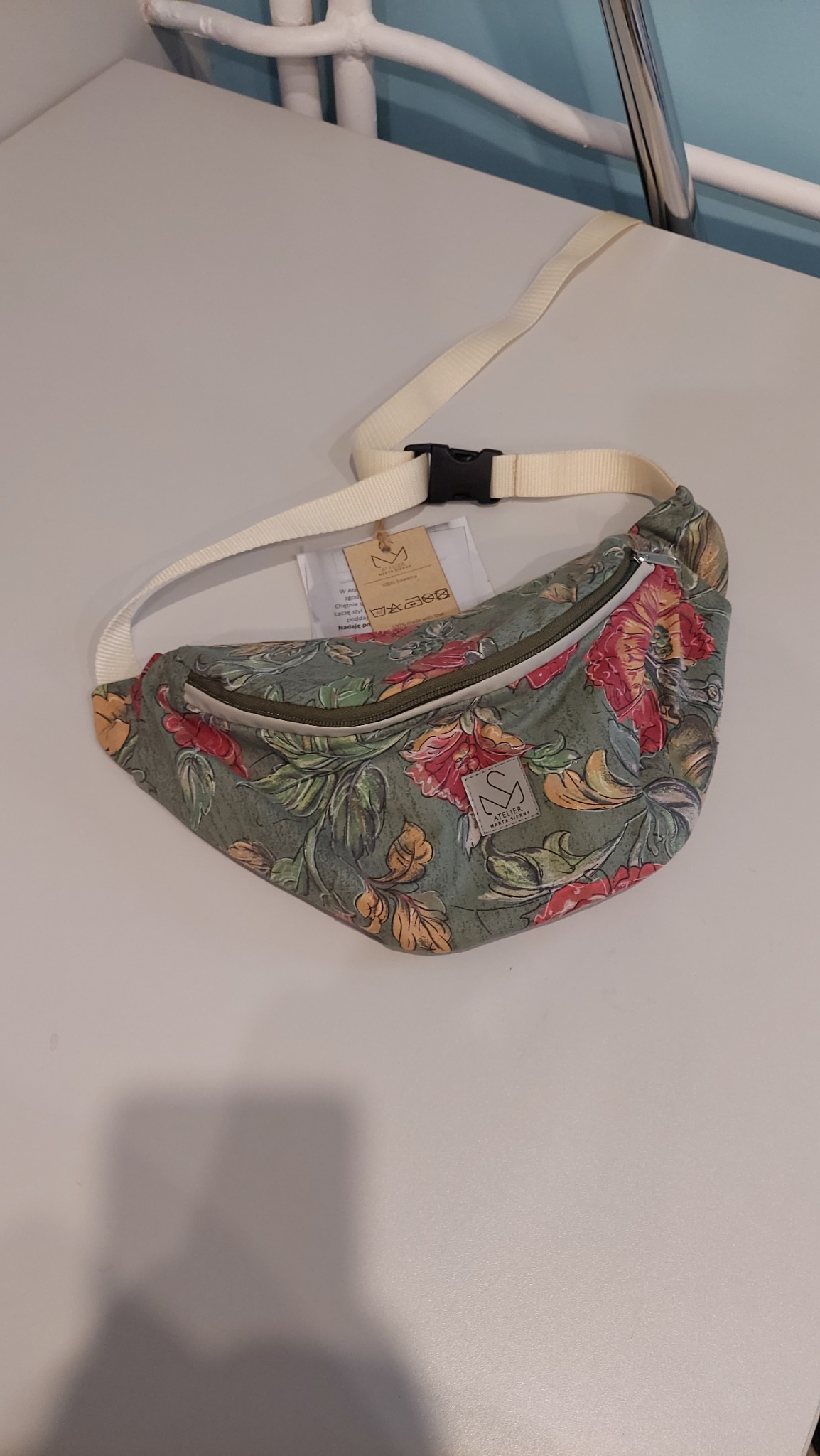 Saszetka nerka z motywem kwiatowym, leżąca na białym blacie. Widoczny pasek z klamrą i metka z logo Atelier Marta Słowy. Unikalny wzór i staranne wykonanie.