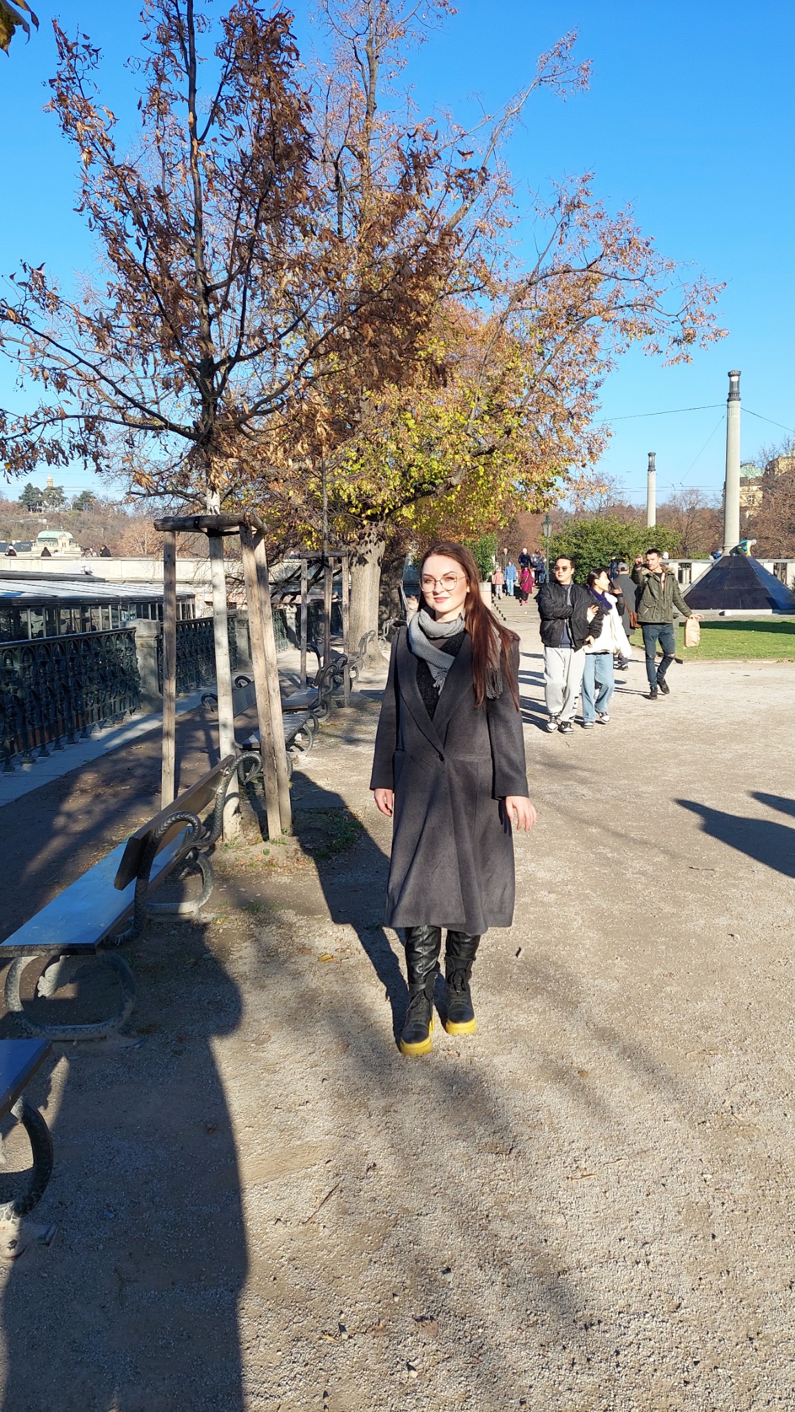 Kobieta w szarym płaszczu i czarnych botkach na tle parku w słoneczny dzień. Jesienne drzewo z brązowymi liśćmi i alejka z ławkami.