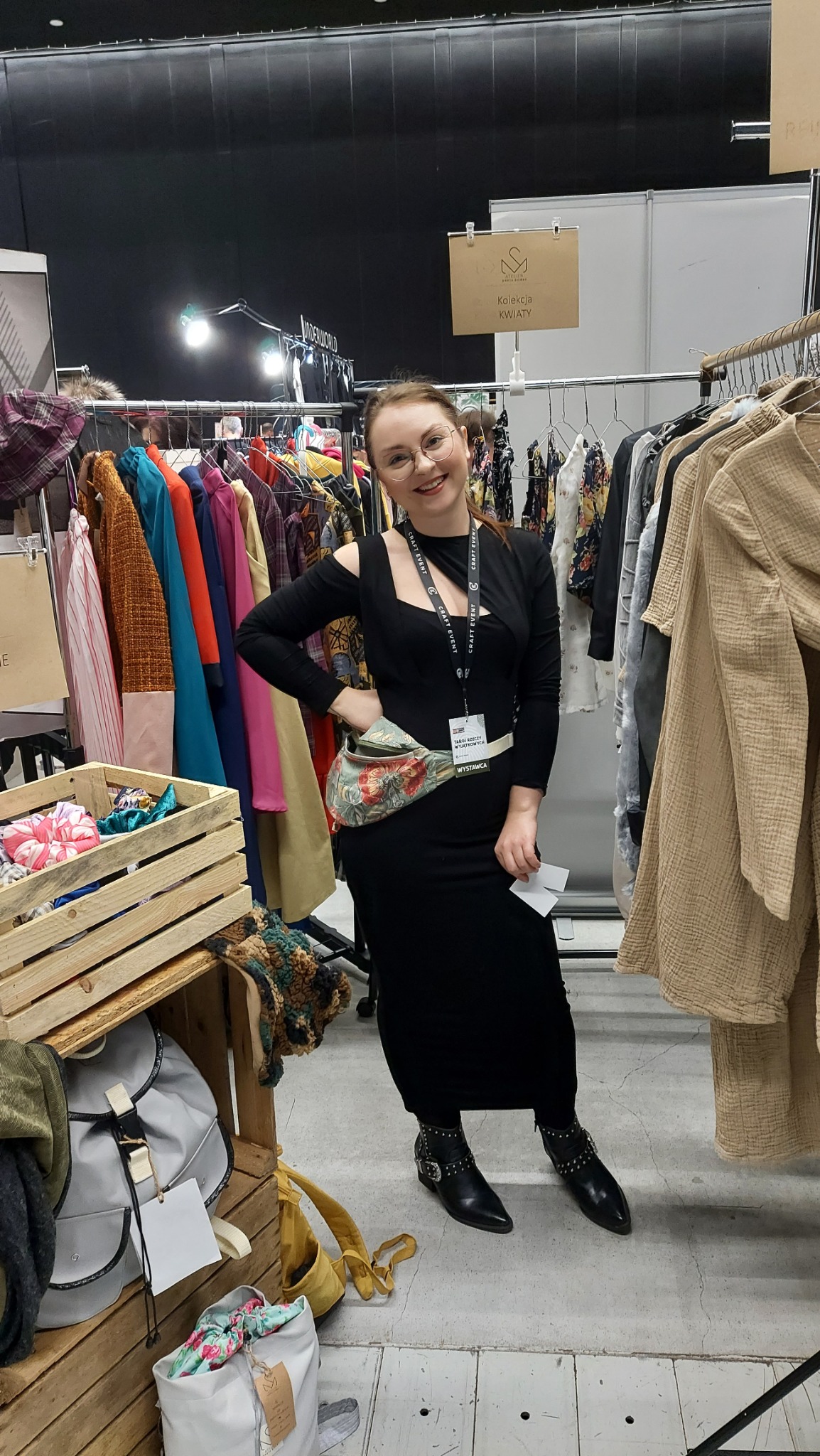 Uśmiechnięta kobieta w czarnej sukience prezentuje się na stoisku z odzieżą, z różnorodnymi ubraniami na wieszakach i drewnianą skrzynią z akcesoriami obok.