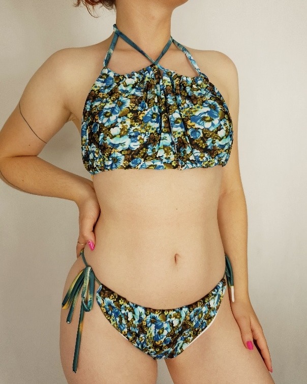 Kobieta prezentuje dwuczęściowy strój kąpielowy z motywem kwiatowym w odcieniach niebieskiego i brązowego. Strój ma wiązania po bokach majtek i na szyi.