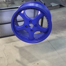 ANL Serwis Tomasz Nawrocki - Niebieska felga aluminiowa zawieszona na łańcuchu w warsztacie, po procesie malowania proszkowego. Widoczne inne elementy metalowe i narzędzia w tle.