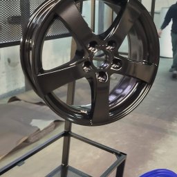 ANL Serwis Tomasz Nawrocki - Błyszcząca, czarna felga samochodowa zawieszona na metalowym stojaku w warsztacie. W tle widoczna niebieska felga i fragment sylwetki osoby.