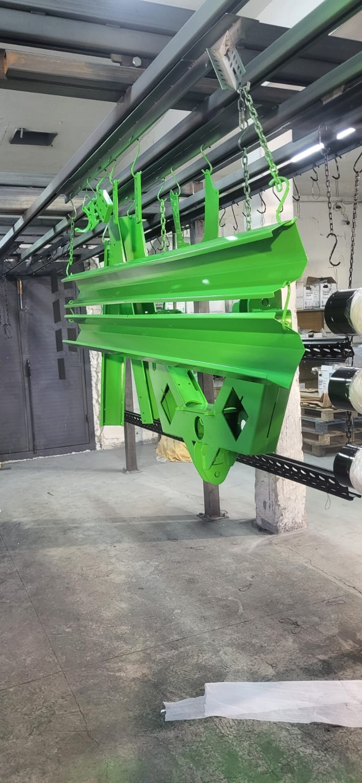 Elementy konstrukcyjne, pomalowane na jaskrawy zielony kolor, wiszą na łańcuchach w hali produkcyjnej. Widoczne ślady po malowaniu na podłodze.