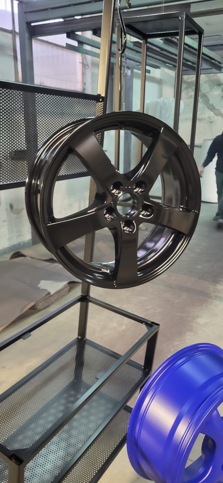 Błyszcząca, czarna felga samochodowa zawieszona na metalowym stojaku w warsztacie. W tle widoczna niebieska felga i fragment sylwetki osoby.