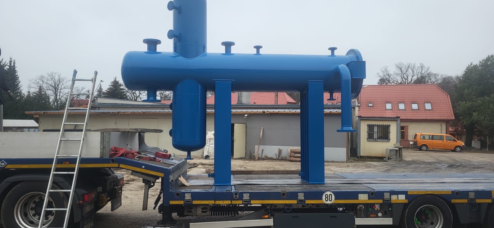 Niebieska, pomalowana konstrukcja stalowa na naczepie ciężarówki. Widoczna drabina, budynki w tle i pochmurne niebo. Elementy konstrukcji spawane.