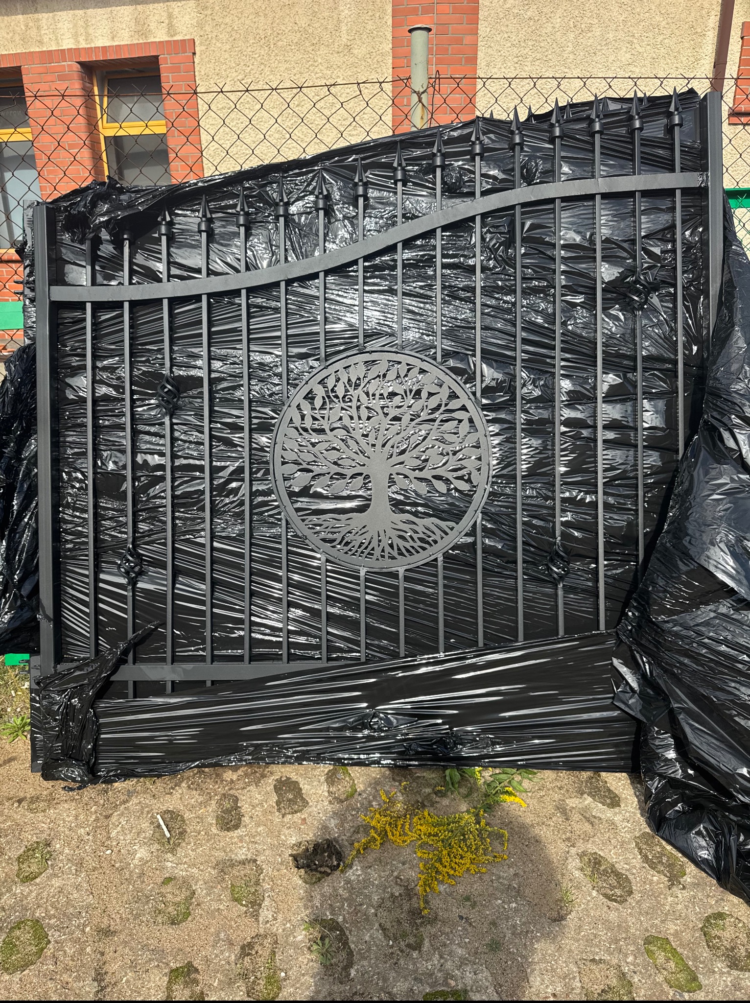 Nowa, szara brama wjazdowa z motywem drzewa, zabezpieczona czarną folią stretch, czeka na montaż. W tle budynek z cegły i siatka ogrodzeniowa.