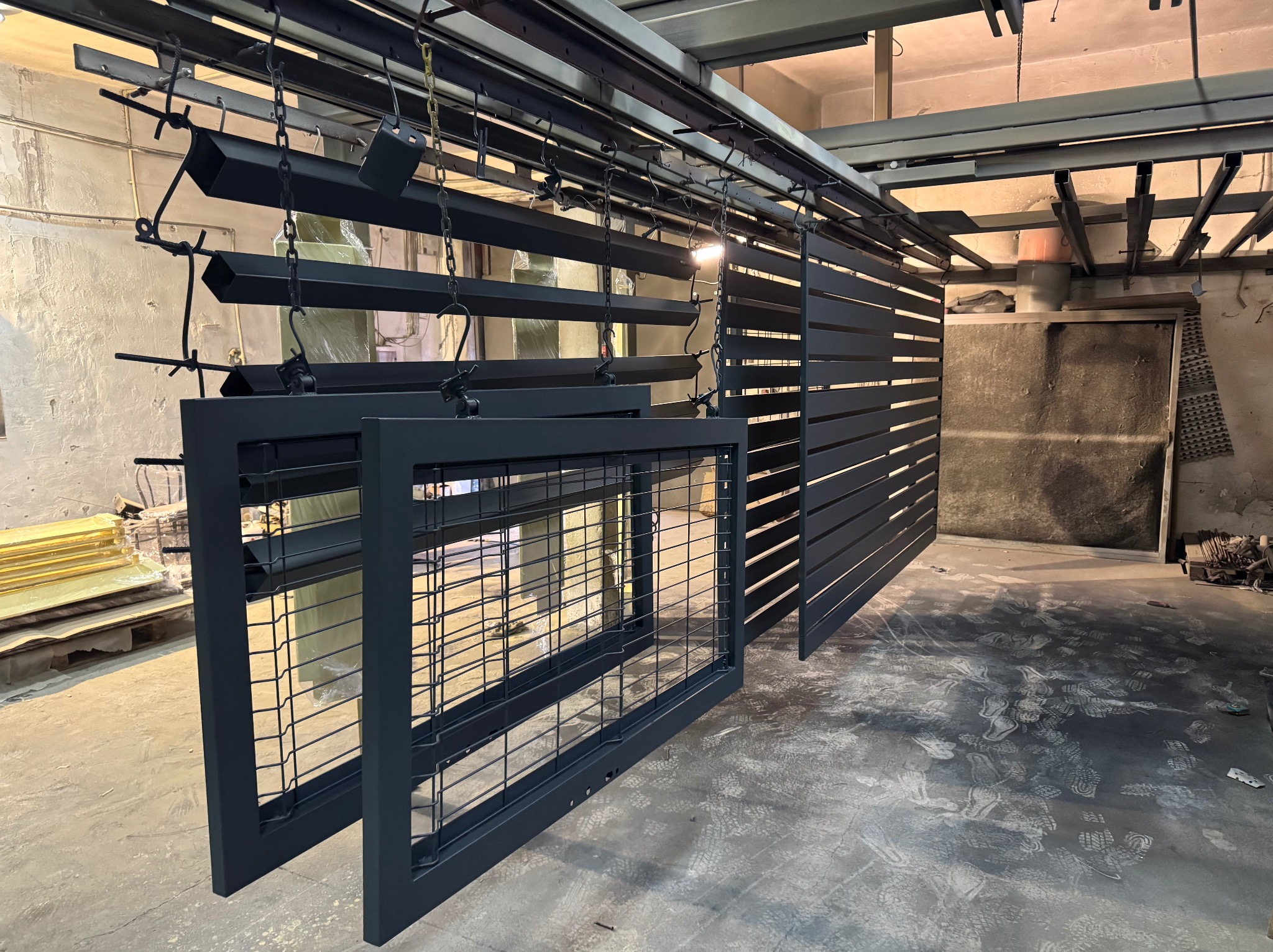 Elementy metalowe (bramy, ogrodzenia) po malowaniu proszkowym, zawieszone na łańcuchach w hali produkcyjnej. Widoczne ślady stóp na podłodze.