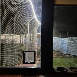 Siatki Ludwik - Zabezpieczony siatką balkon z drzwiczkami dla kota, widok z wnętrza. Siatka tworzy tunel na zewnątrz, zapewniając bezpieczeństwo zwierzęciu. Widoczne miski z jedzeniem.