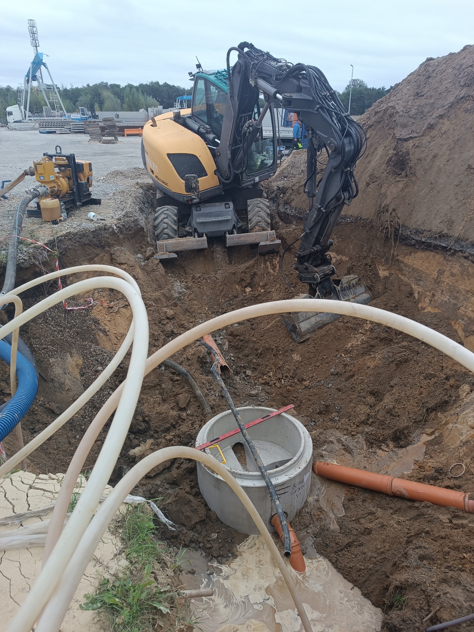 Żółta koparka Mecalac w wykopie z rurami i betonowym kręgiem. Widoczna pompa odwadniająca. W tle wesołe miasteczko i hałda ziemi.