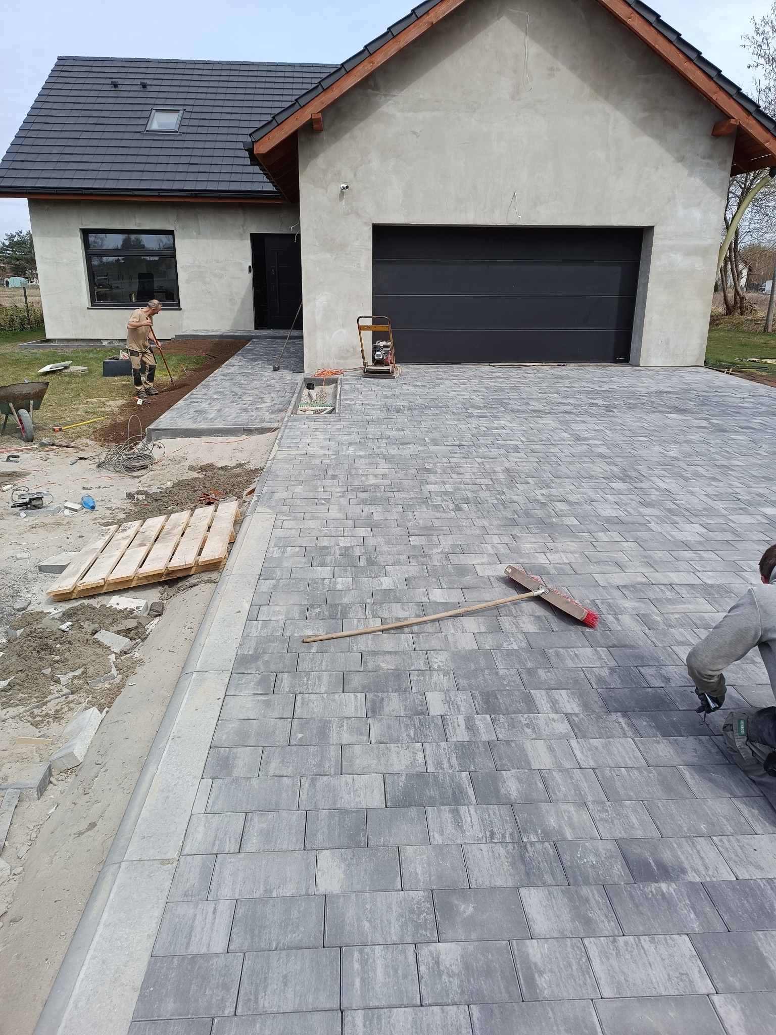 Układanie szarej kostki brukowej na podjeździe do domu z czarną bramą garażową. W tle widoczny pracownik z narzędziem. Częściowo ukończona realizacja.