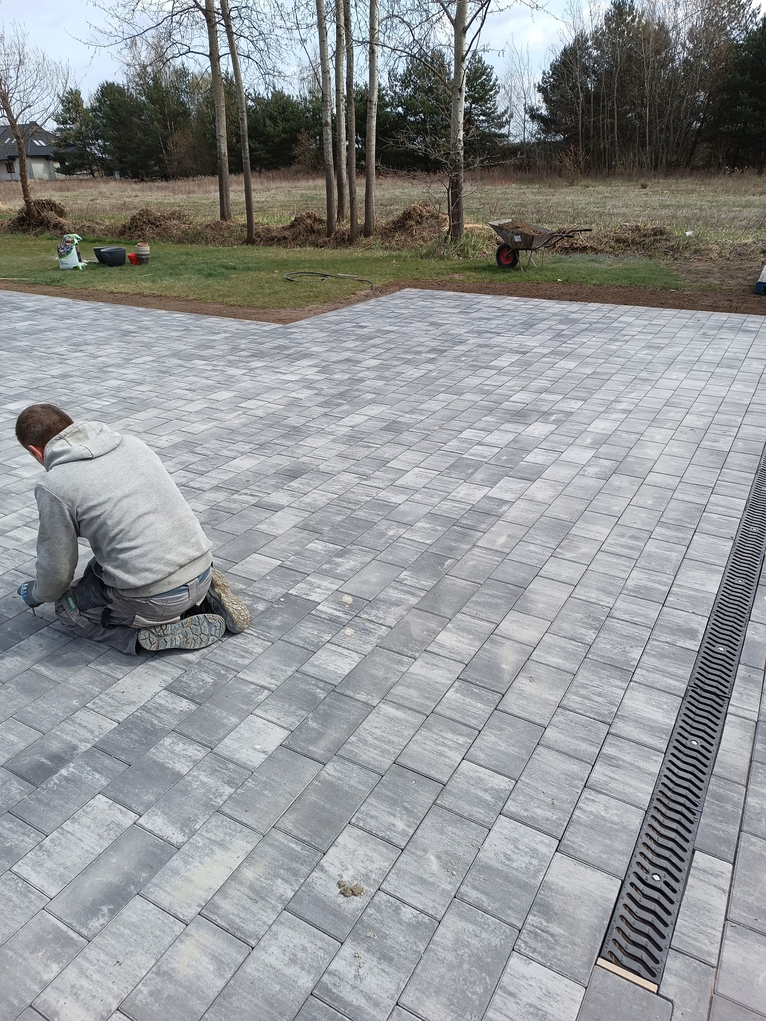Pracownik na kolanach dopasowuje szarą kostkę brukową z odpływem liniowym. W tle drzewa i krajobraz wiejski. Widoczny fragment ułożonej nawierzchni.