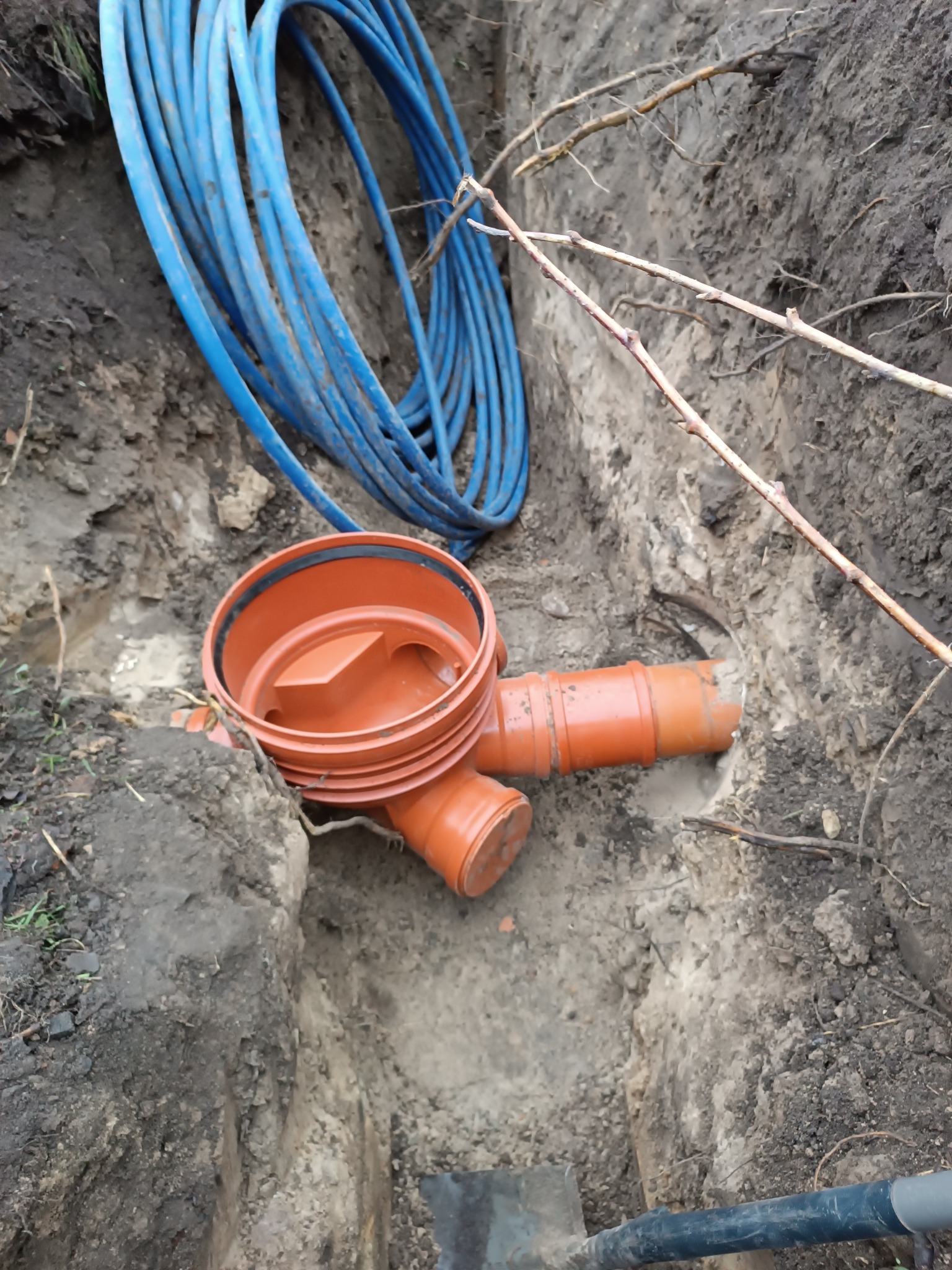 Pomarańczowy trójnik kanalizacyjny w wykopie ziemnym z niebieskimi rurami i fragmentem łopaty w dolnym rogu. Widoczne gałęzie i ziemia.