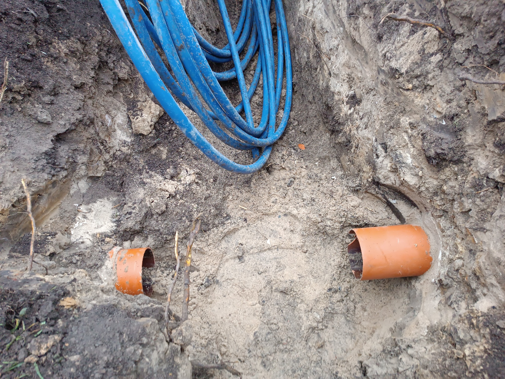 Wykop z widocznymi rurami kanalizacyjnymi i niebieskimi kablami. Prace ziemne w trakcie realizacji, widoczne detale wykopu i ułożonych elementów instalacji.