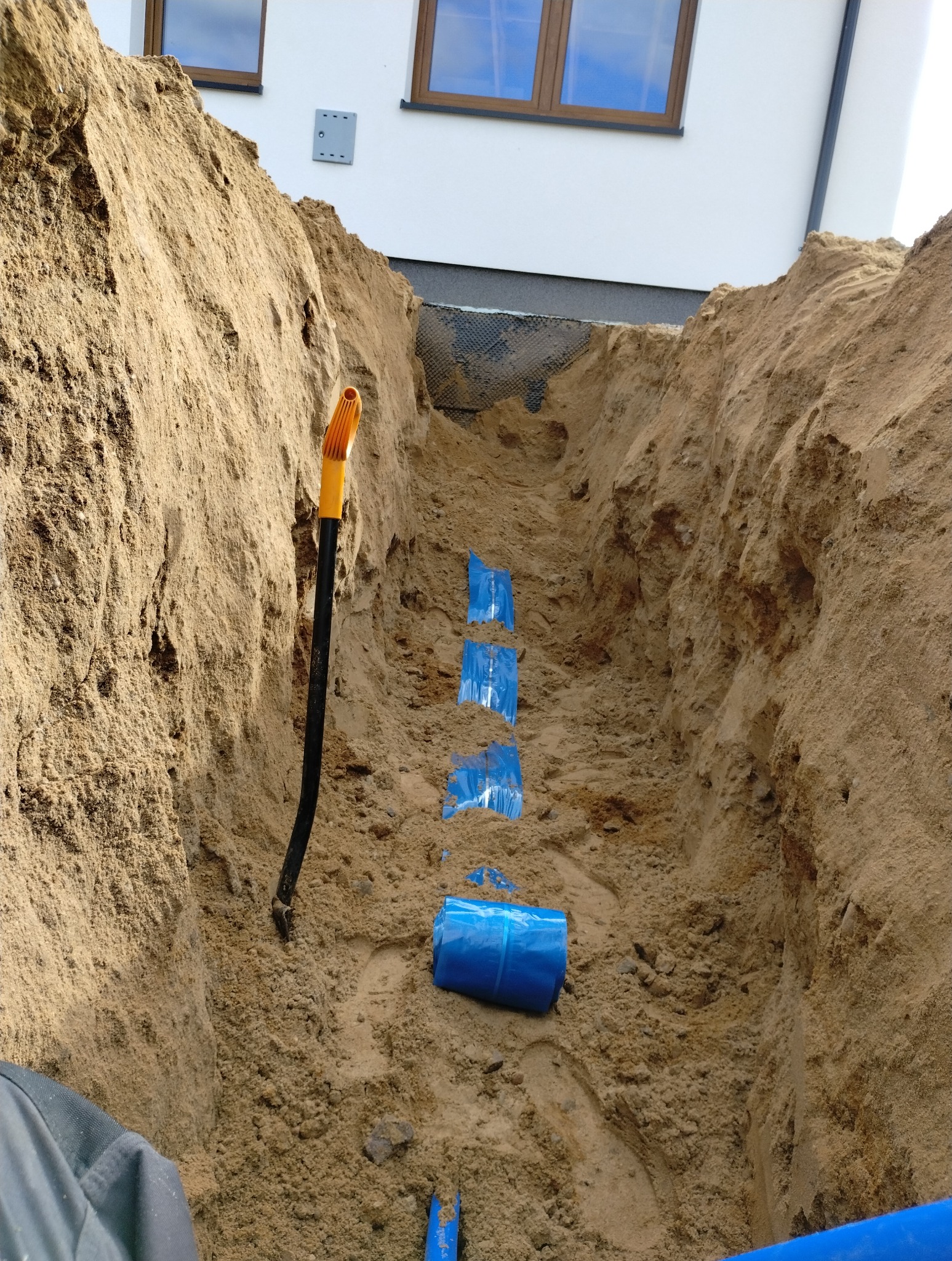 Wykop w piasku z rurami owiniętymi niebieską folią, widoczna łopata. Instalacja w ziemi przed budynkiem, etap prac ziemnych.