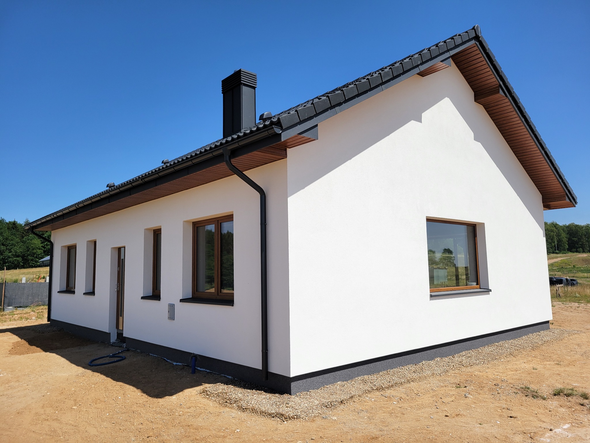 Biały dom jednorodzinny z czarnym dachem i orynnowaniem, brązowe ramy okien, widok z zewnątrz w słoneczny dzień, świeżo wykończona elewacja.