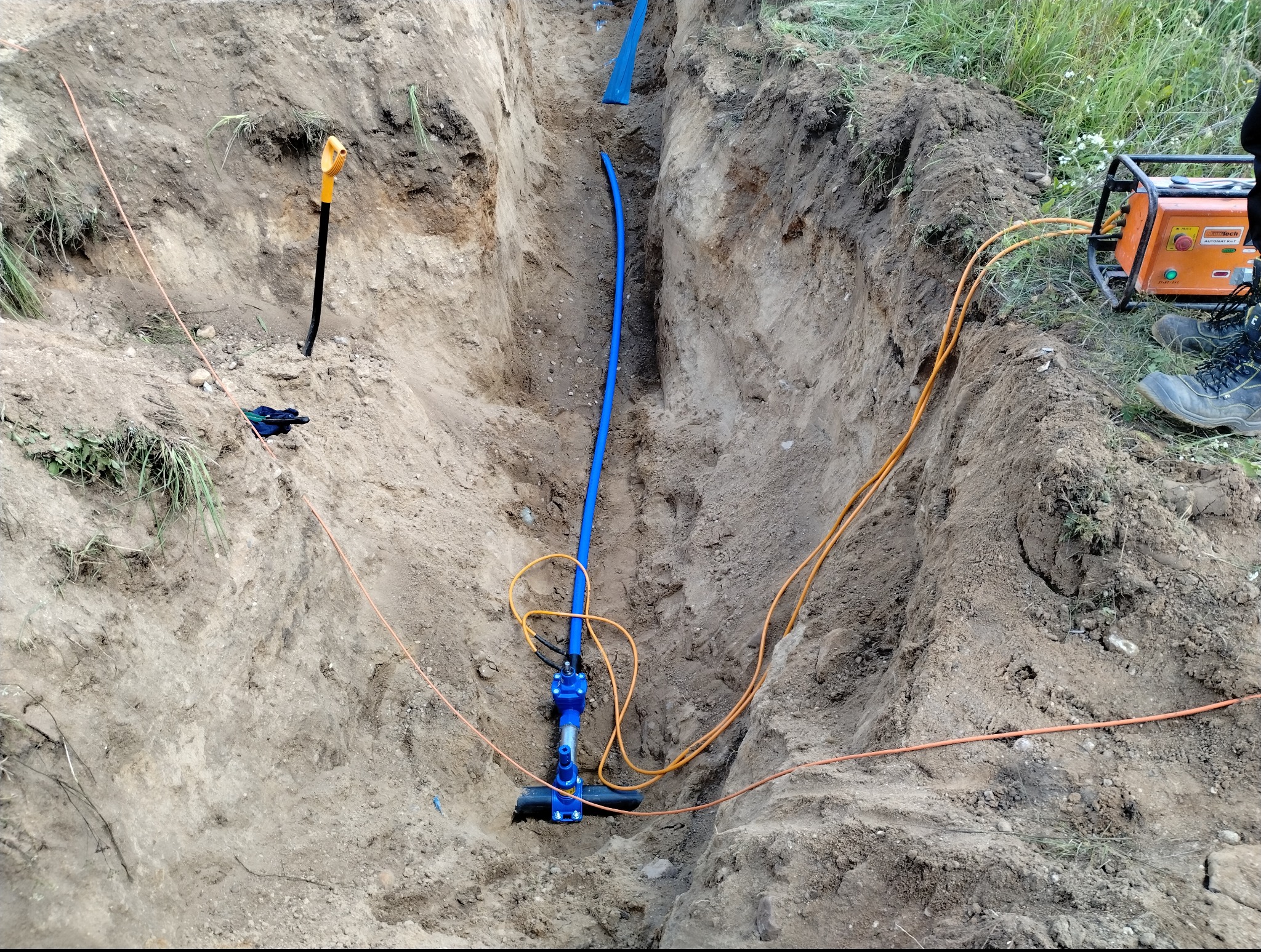 Wykop z niebieską rurą, prawdopodobnie instalacja podziemna w trakcie montażu. Widoczne narzędzia i przewody. Ziemny krajobraz, przygotowania do zasypania.