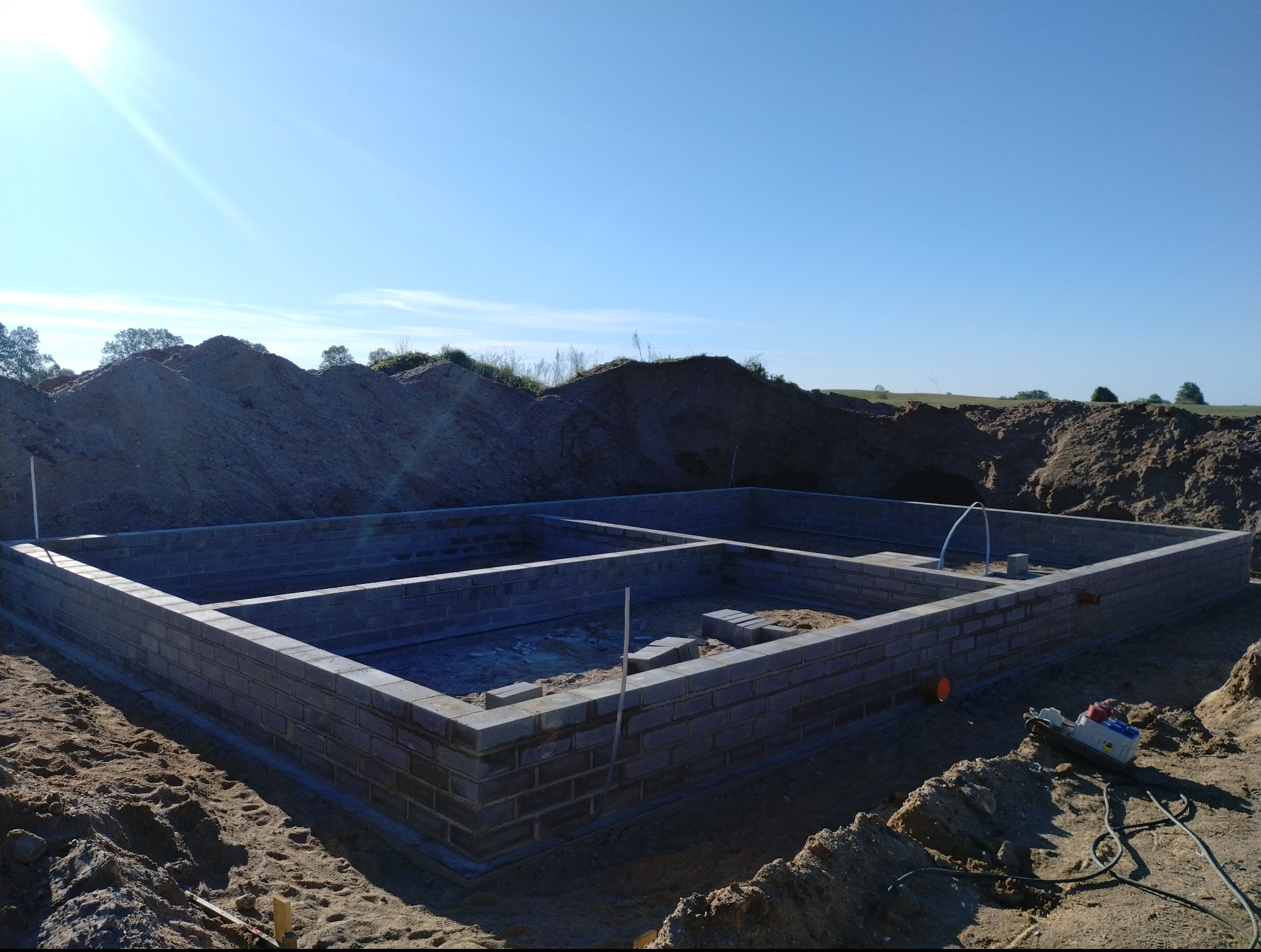 Wymurowane fundamenty domu z bloczków betonowych na tle wykopu i błękitnego nieba. Widoczny sprzęt budowlany. Etap budowy w słoneczny dzień.