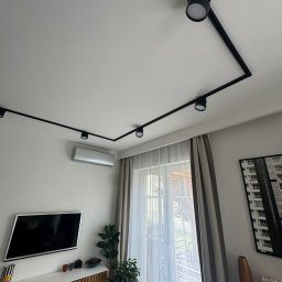 Remo-Bud - Nowoczesny salon z czarnym systemem szynowym oświetlenia sufitowego, klimatyzacją i telewizorem. Minimalistyczny design wnętrza z roślinami i dekoracjami.