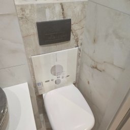 Remo-Bud - Kompakt WC z podtynkową spłuczką Geberit, obudowany płytkami imitującymi marmur. Widoczny dozownik mydła i rolka papieru toaletowego. Wykończenie łazienki w odcieniach beżu.