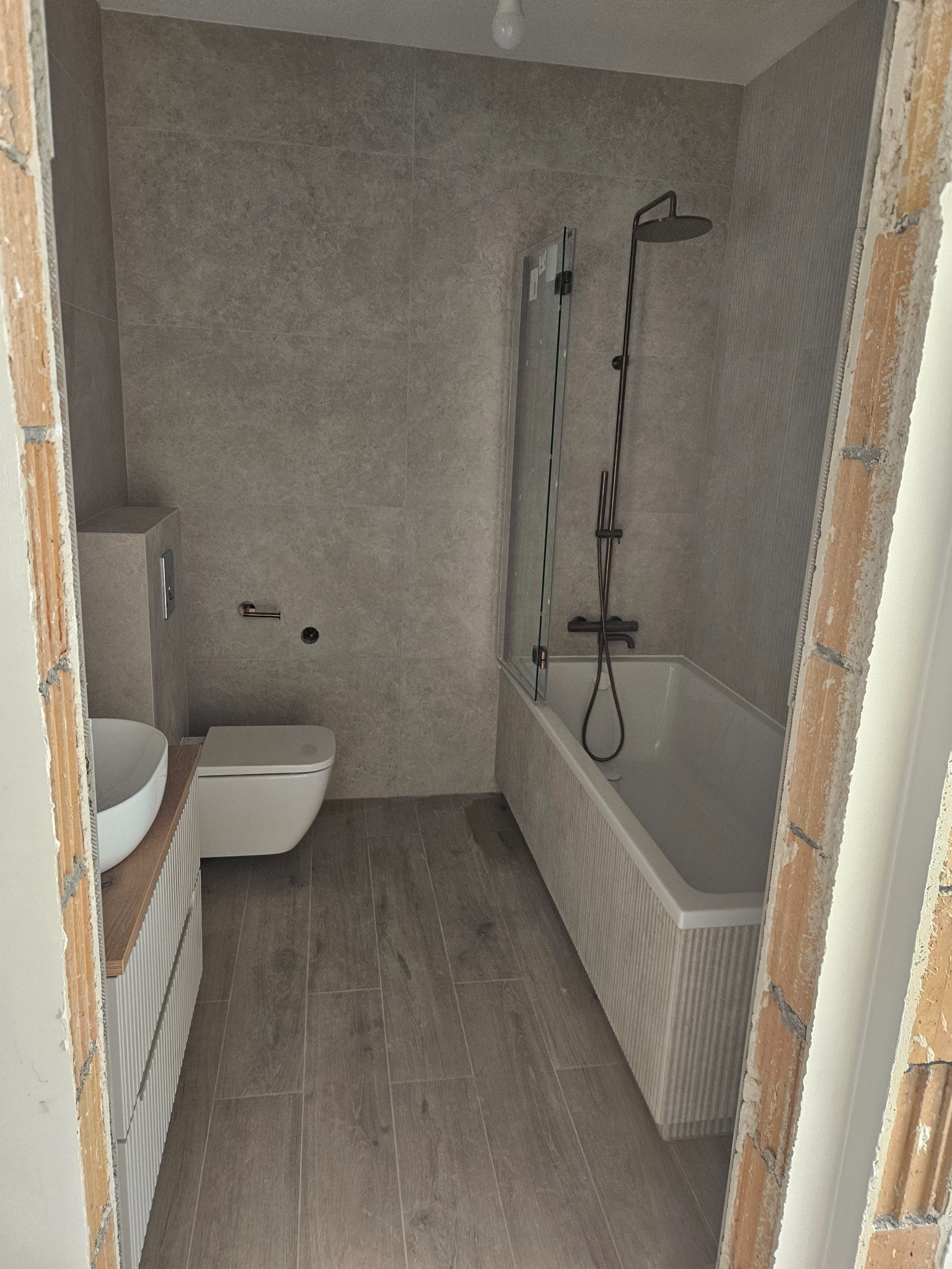 Nowoczesna łazienka w szarościach z wanną, kabiną prysznicową, umywalką i toaletą. Ukończone płytki na ścianach i podłodze. Widok z perspektywy wejścia.