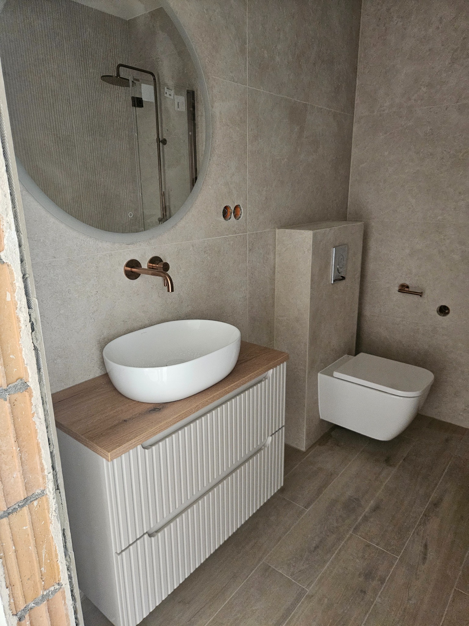 Ukończona łazienka w beżowej tonacji z owalnym lustrem, białą umywalką nablatową i podwieszaną toaletą. Widoczne płytki imitujące drewno na podłodze.