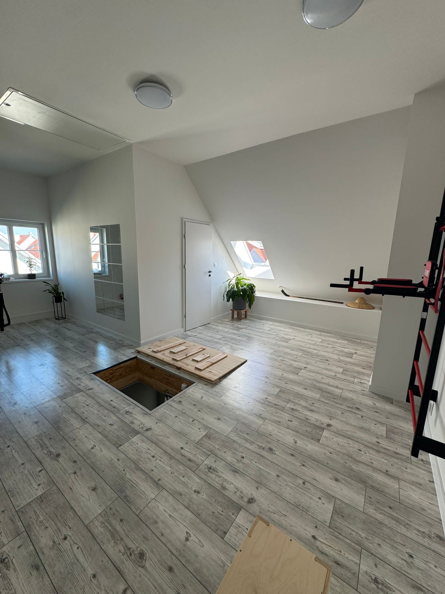 Jasne poddasze z panelami podłogowymi imitującymi drewno. Otwór w podłodze zabezpieczony drewnianymi deskami. Ściany białe, okna, roślina doniczkowa.