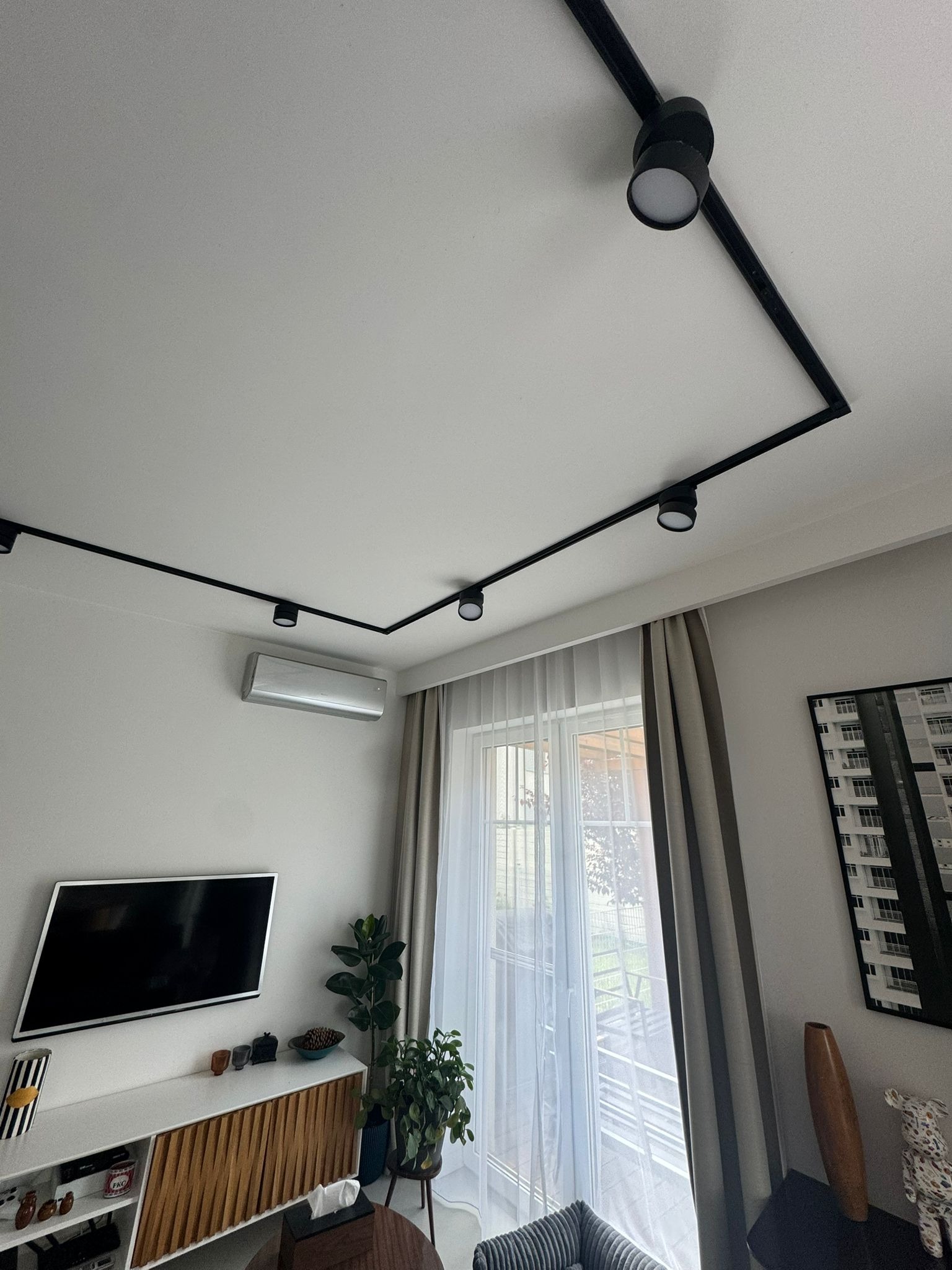 Nowoczesny salon z czarnym systemem szynowym oświetlenia sufitowego, klimatyzacją i telewizorem. Minimalistyczny design wnętrza z roślinami i dekoracjami.