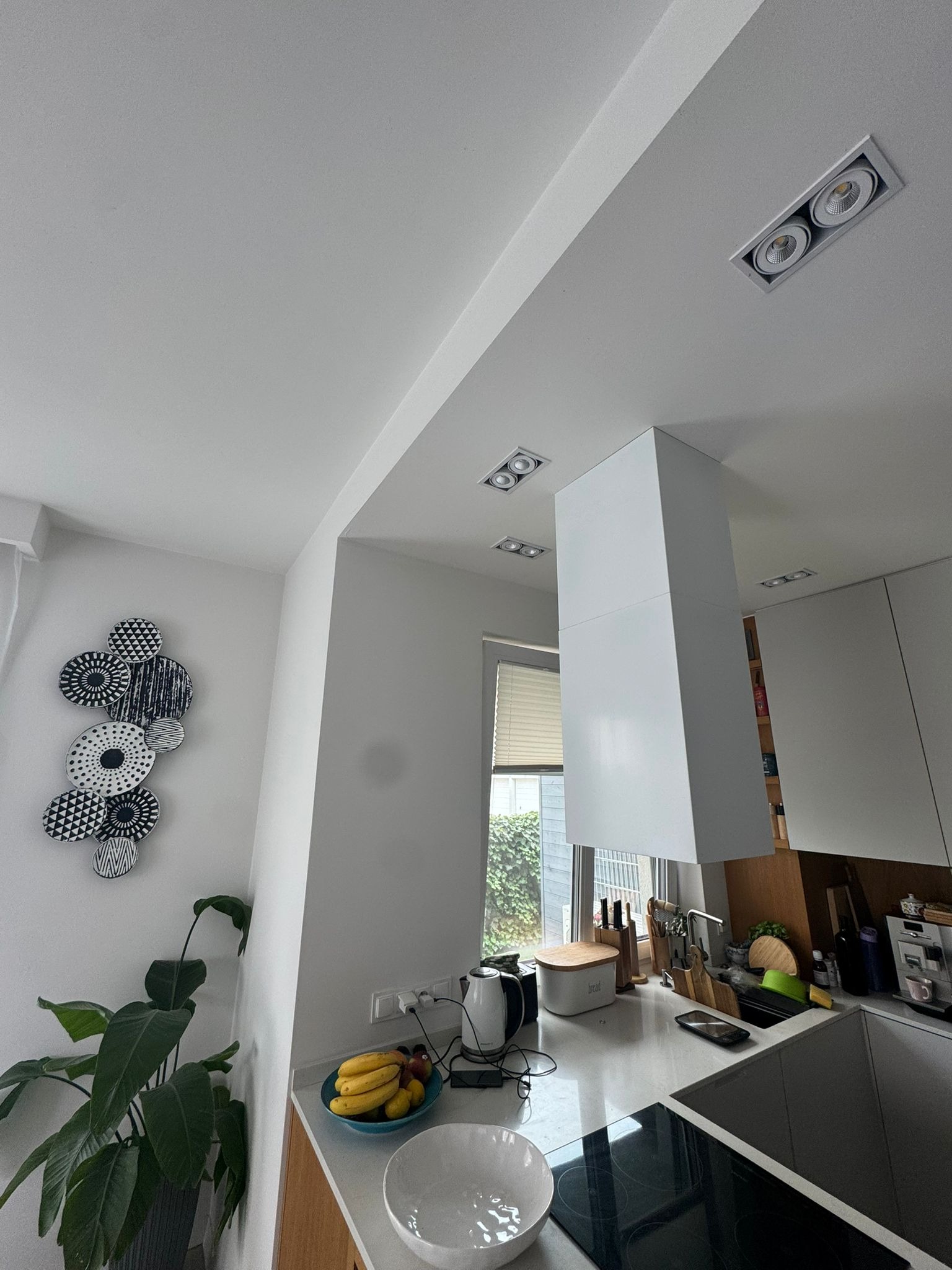 Nowoczesna, jasna kuchnia z białymi meblami, okapem i oświetleniem punktowym. Na ścianie dekoracja z talerzy, a na blacie owoce i akcesoria kuchenne.