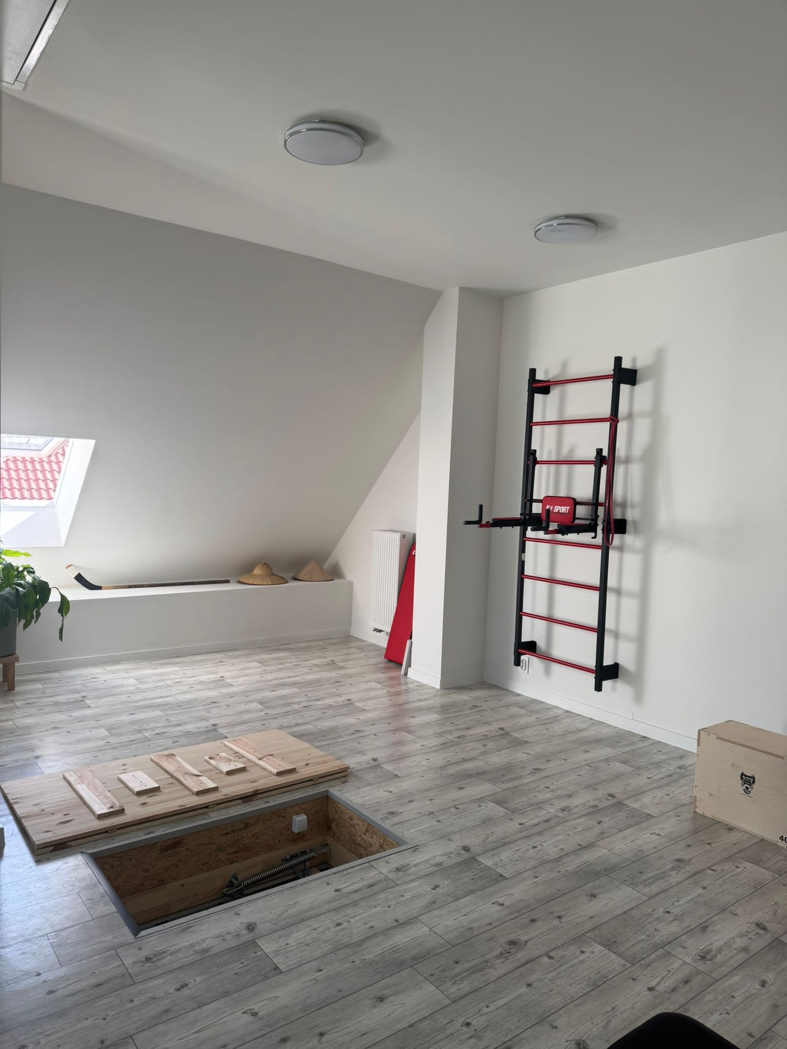 Jasne poddasze z drewnianą podłogą, otworem w podłodze z widocznym mechanizmem, drabinką gimnastyczną na ścianie i oknem dachowym.